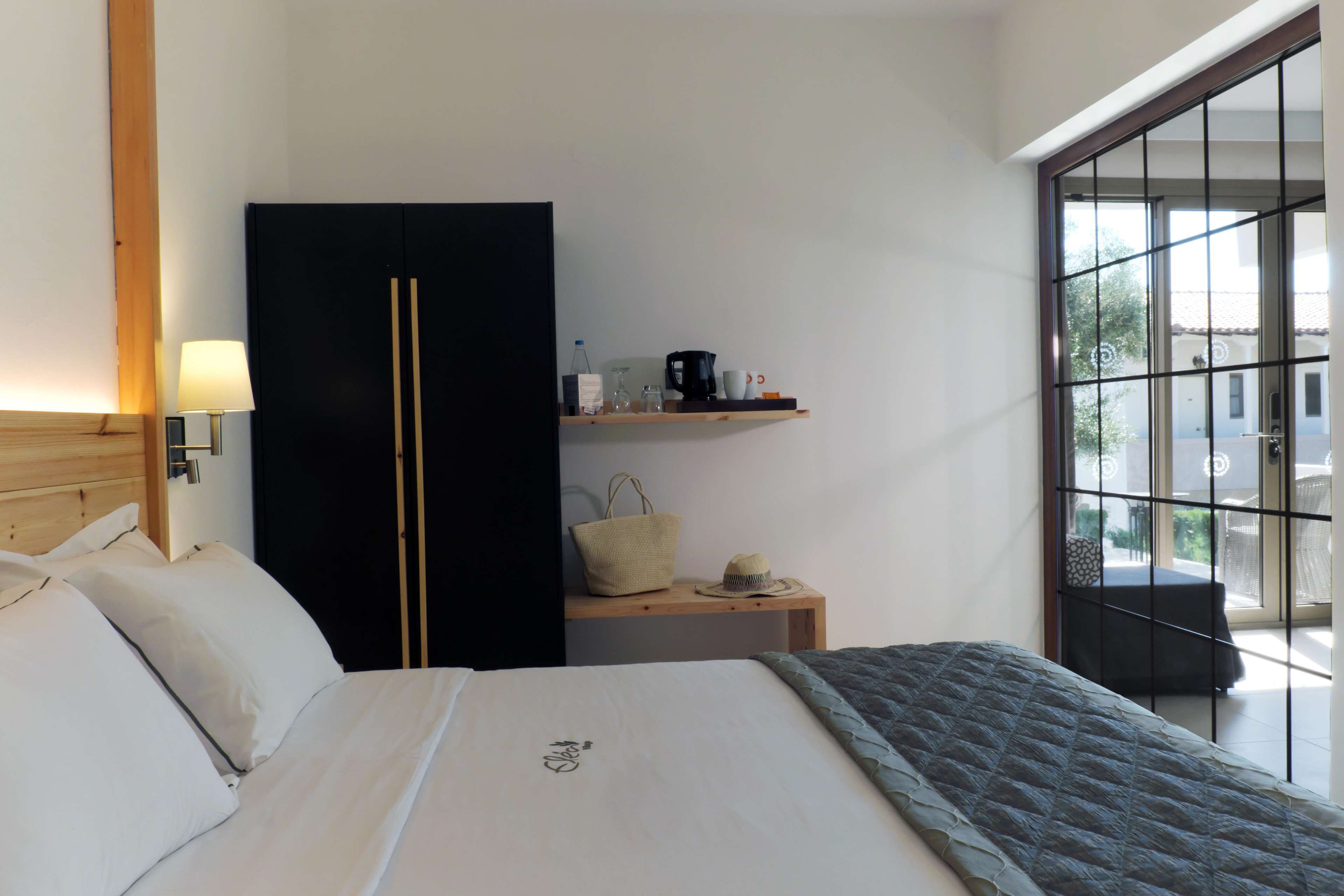 Habitación de hotel con una cama, un armario, una mesita de noche con lámpara, un estante con artículos de tocador y vistas a través de una ventana.