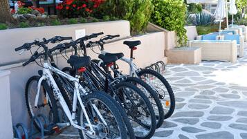 A row of bicycles parked against a wall in an outdoor seating area with stone flooring and greenery in the background.