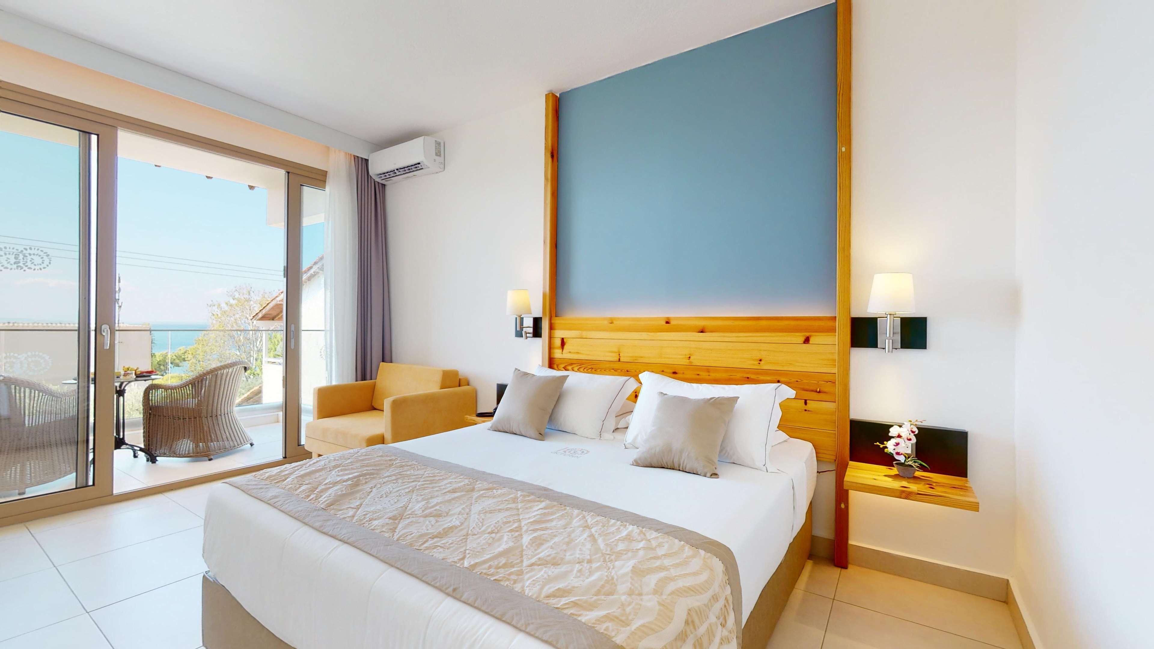 A hotel room with a large bed, a wooden headboard, two bedside tables with lamps, a view of the outdoors through a sliding glass door, and a balcony with wicker furniture.