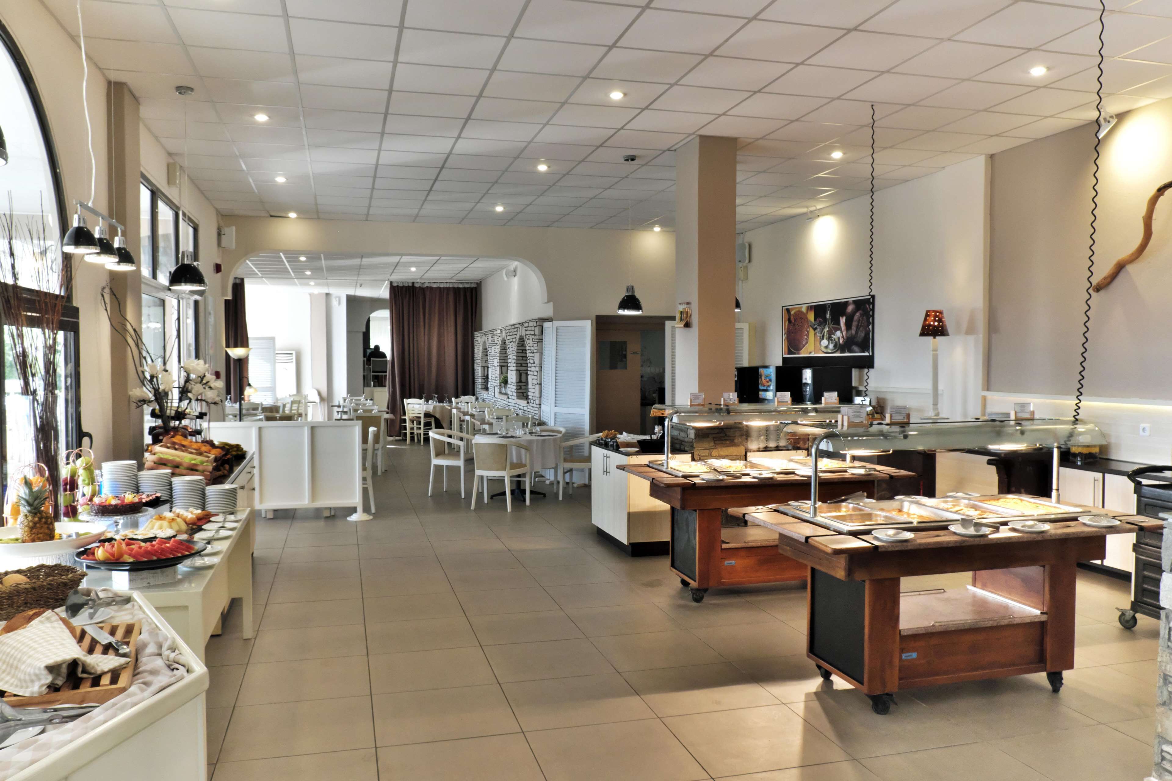 A spacious hotel dining area with a buffet setup, multiple seating arrangements, and a kitchen in the background.