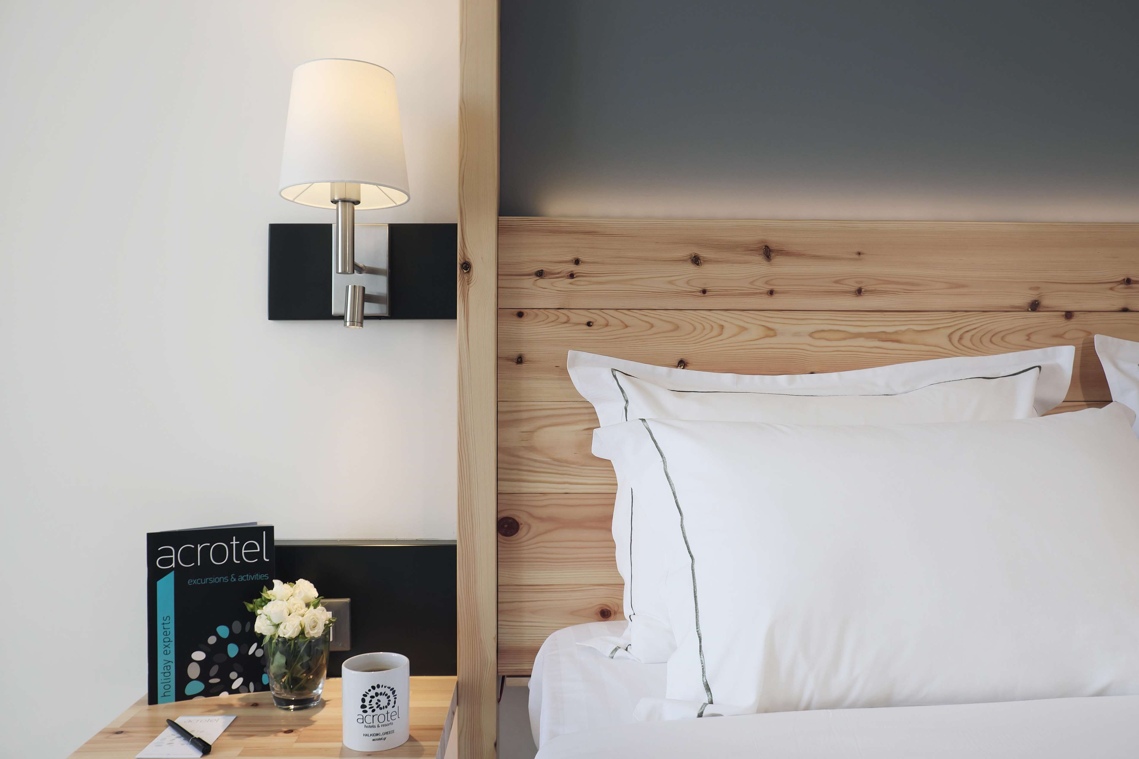 A hotel room with a wooden headboard, a bedside lamp, a nightstand with a brochure, a vase with flowers, and a cup.