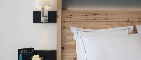 A hotel room with a wooden headboard, a bedside lamp, a nightstand with a brochure, a vase with flowers, and a cup.