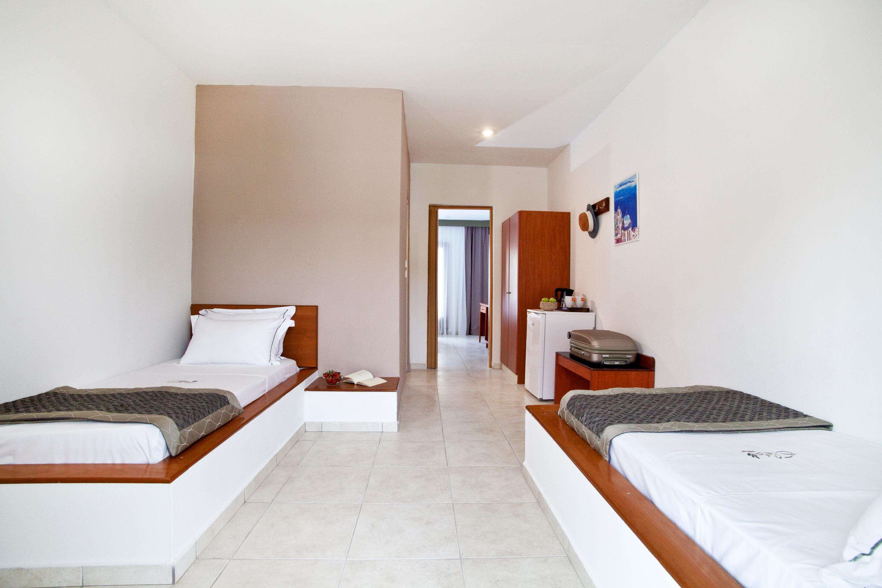 A hotel room with two single beds, a small desk, a refrigerator, and a door leading to another room.