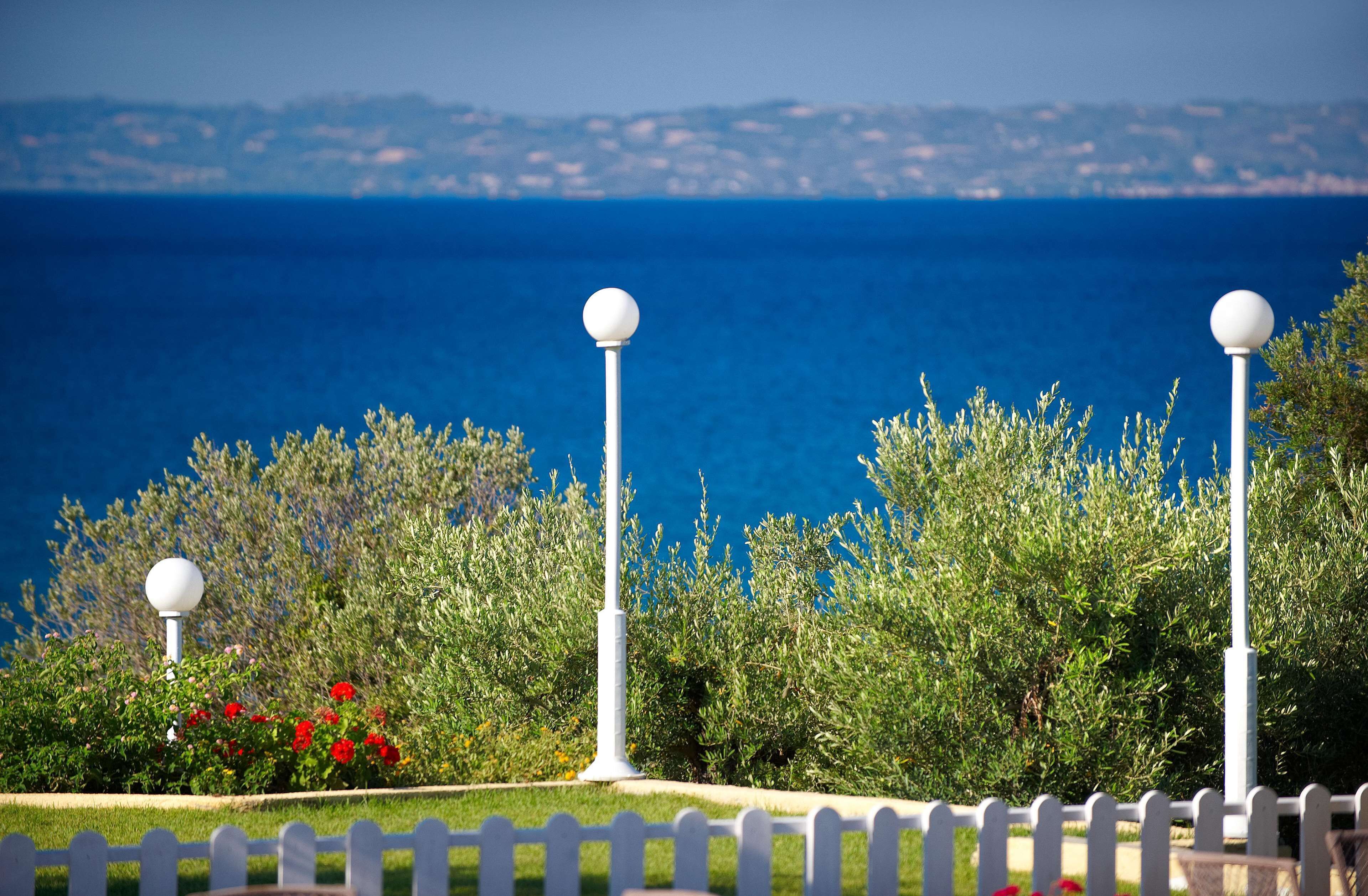 Vista costera con valla de piquete blanca, follaje verde y mar azul claro.