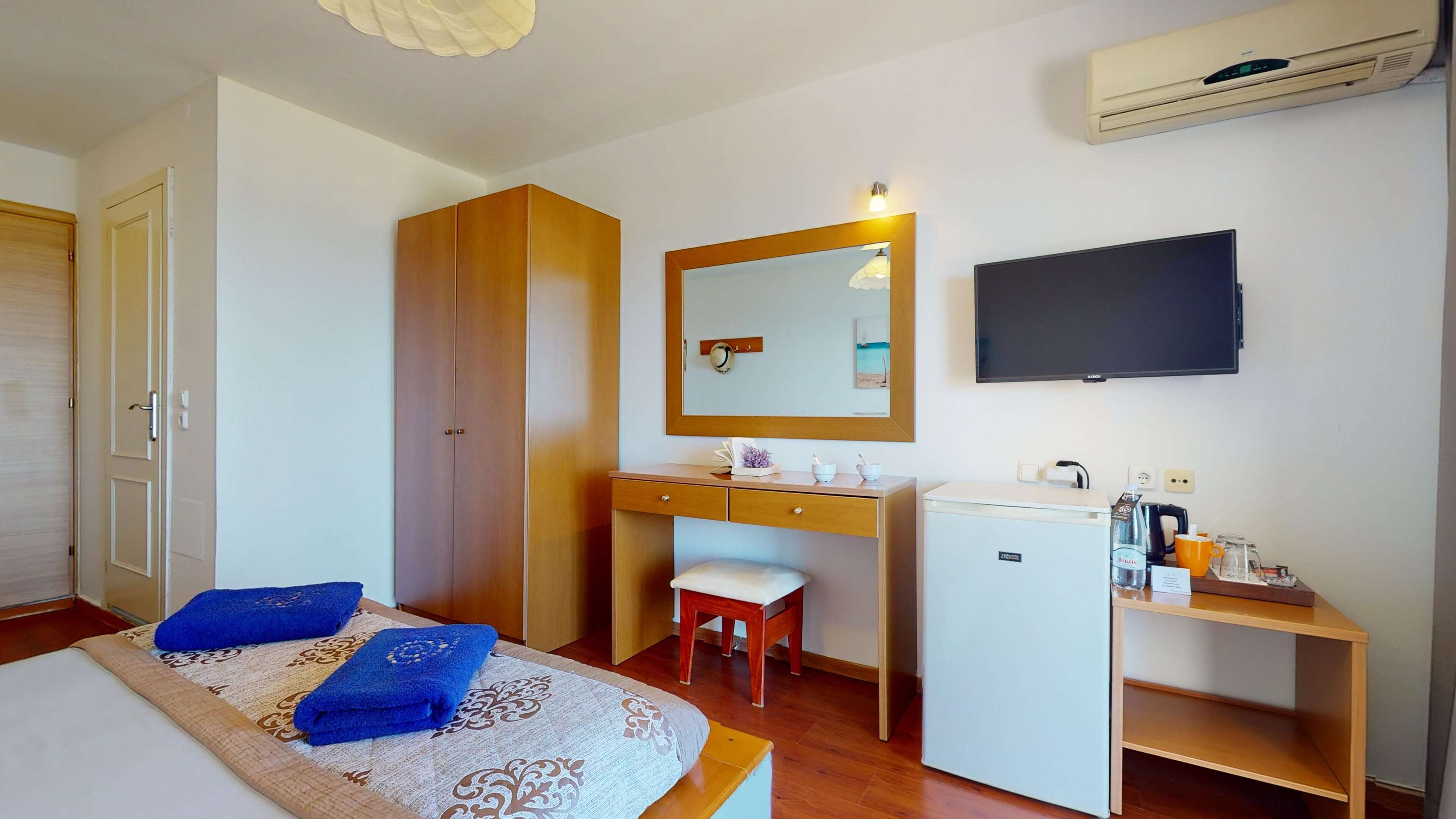 Quarto com cama, escrivaninha, cadeira, pequena geladeira, televisão e ar-condicionado.