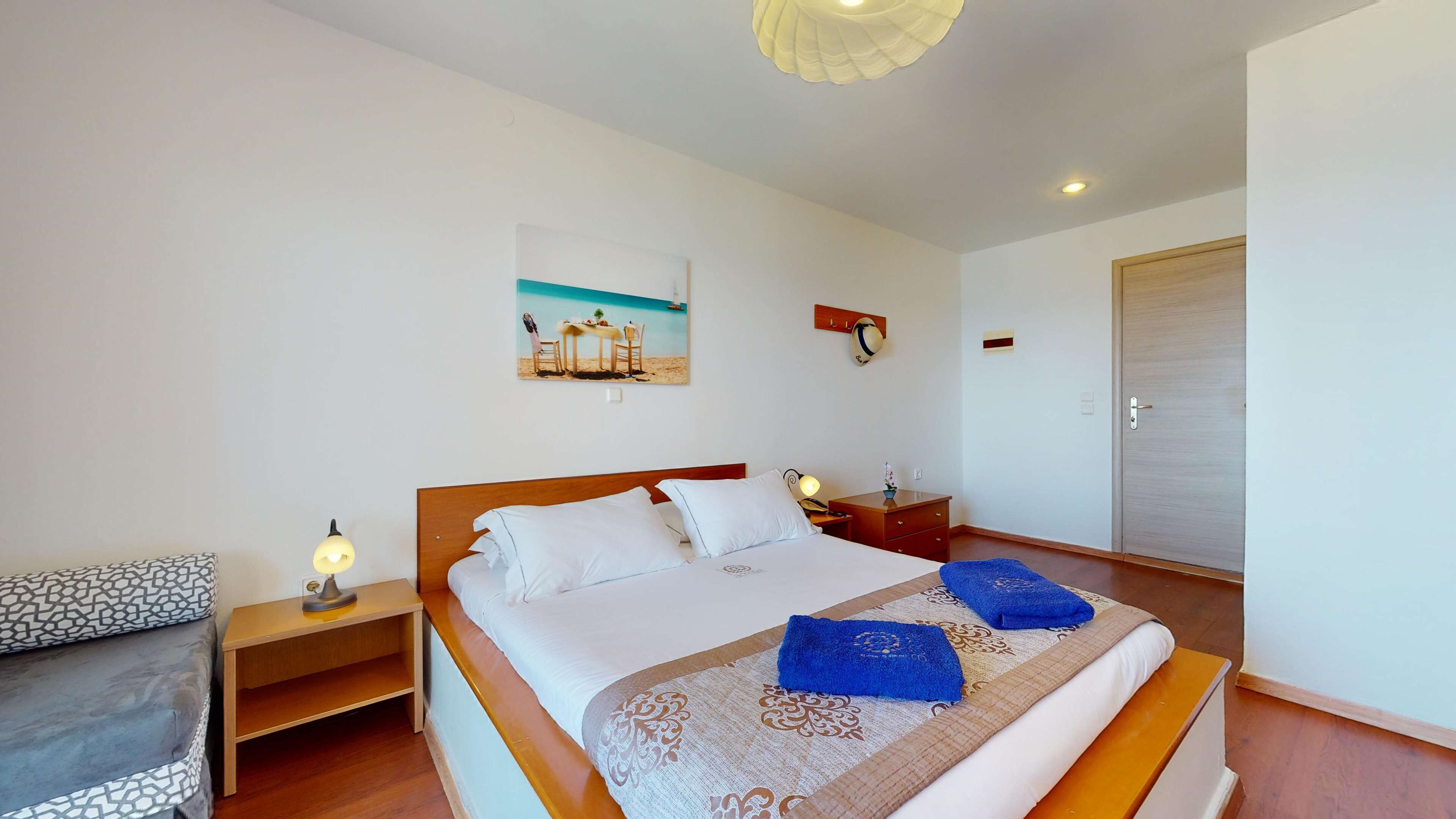 A hotel room with a bed, a nightstand, a lamp, a painting of a beach scene, a wooden door, and a small table with a plant.