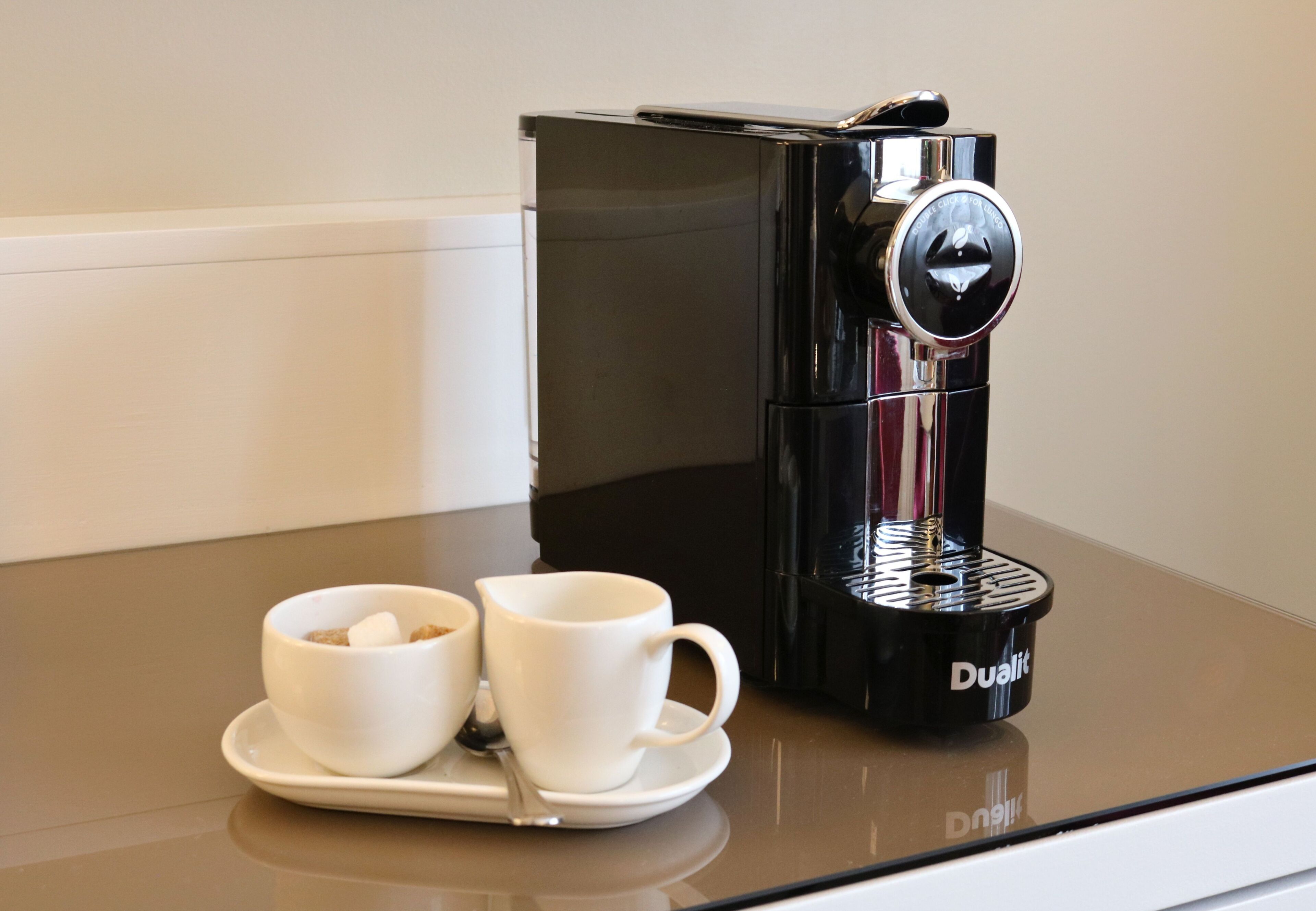 Una cafetera negra con la marca 'Dualit' sobre una superficie reflectante.