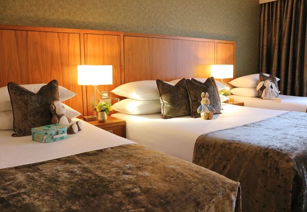 Habitación de hotel con dos camas, cabecero de madera, lámparas de noche, almohadas, peluches y una mesita de noche con un jarrón de flores.
