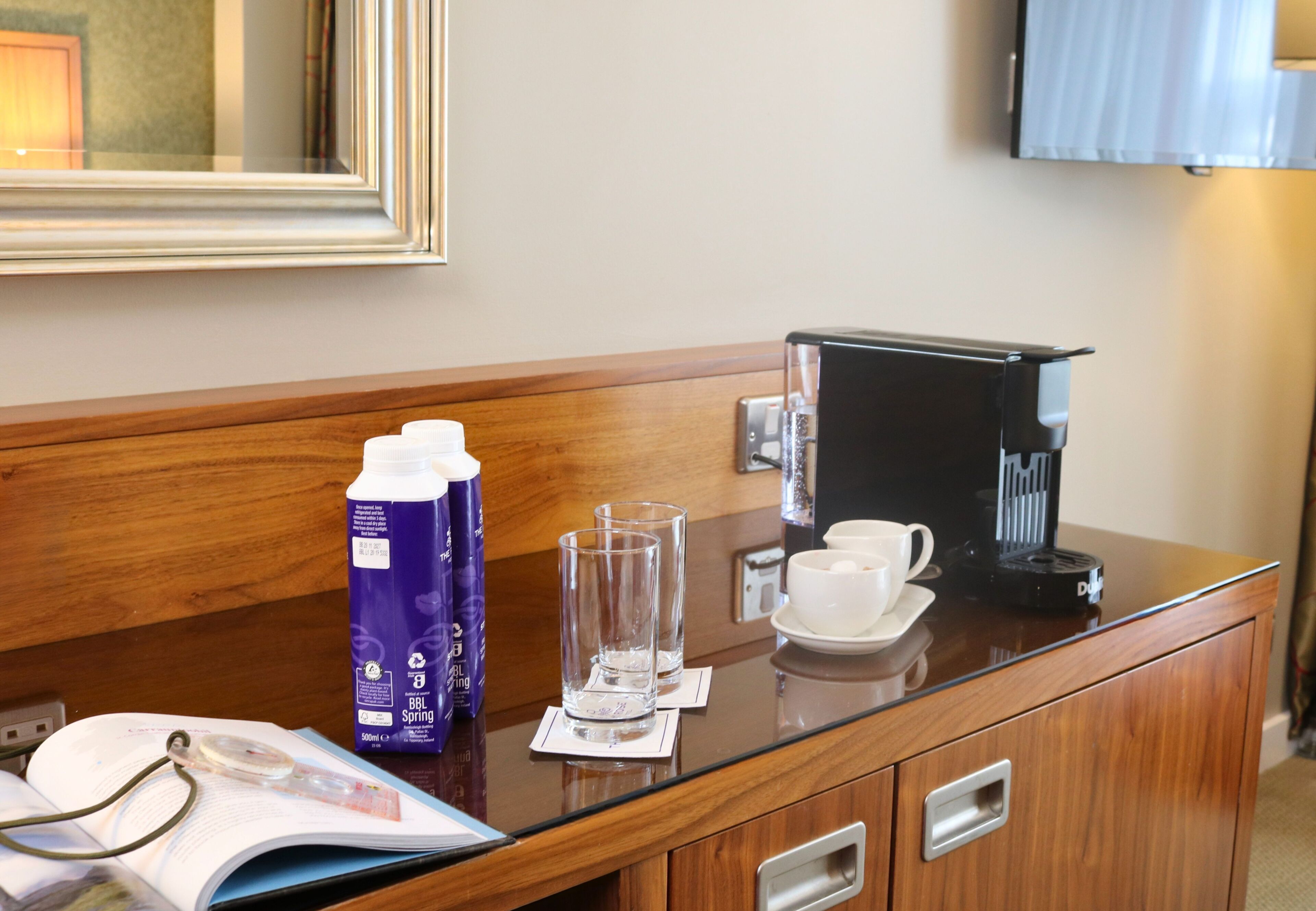 A hotel room with a wooden desk, a coffee maker, a bottle of BBL Spring water, two glasses, a cup, and a book.