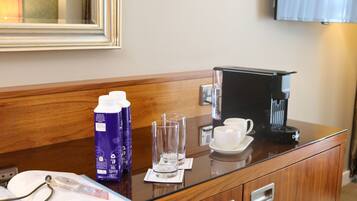A hotel room with a wooden desk, a coffee maker, a bottle of BBL Spring water, two glasses, a cup, and a book.