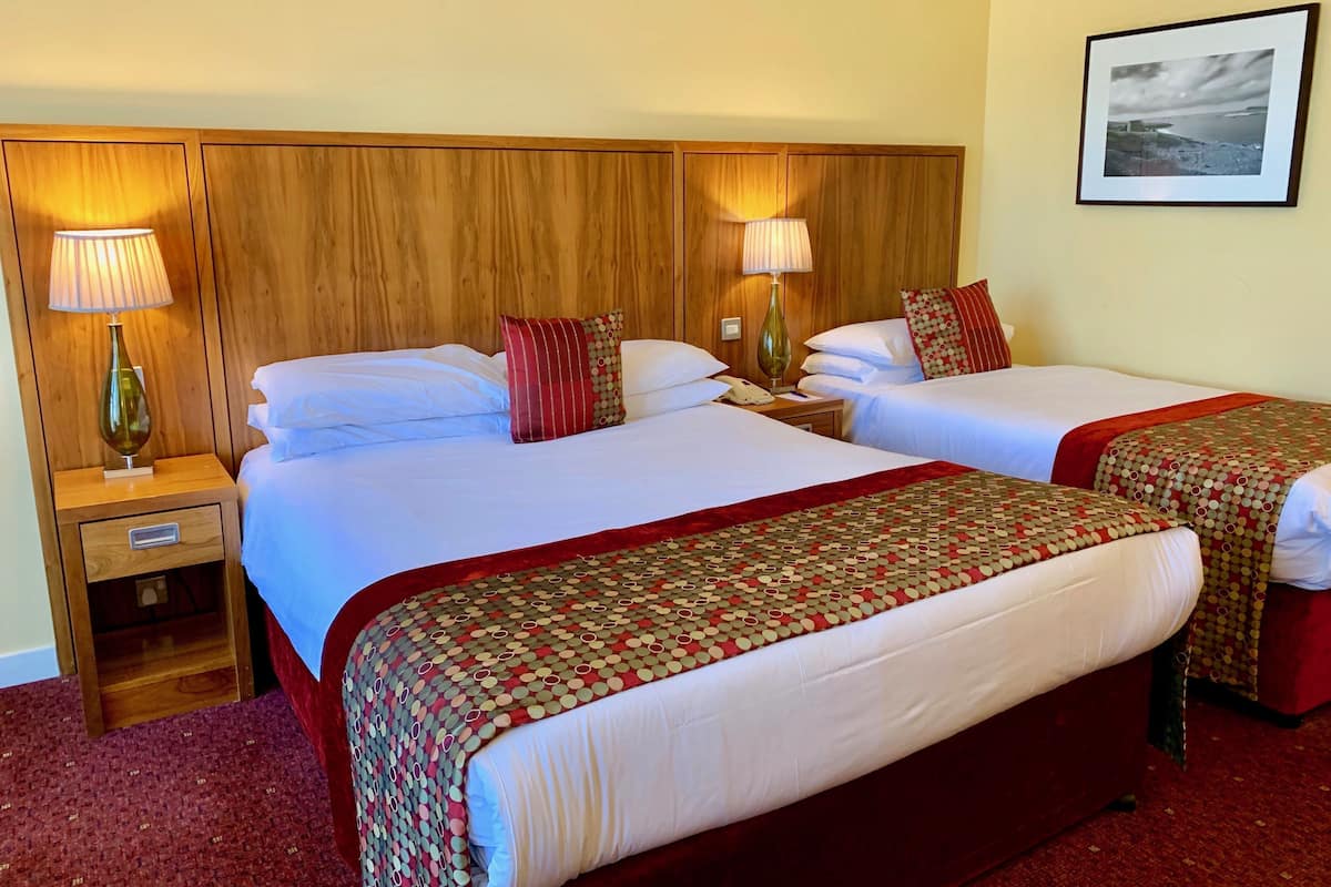 Habitación de hotel con dos camas, cabecero de madera, lámparas de noche, mesita de noche y un cuadro en la pared.