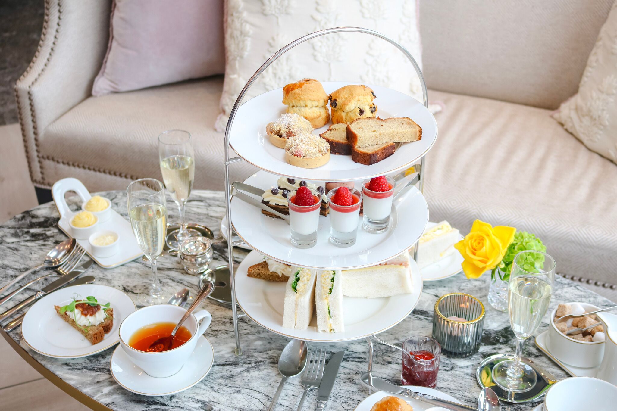 A three-tiered tea service tray with sandwiches, scones, and desserts, accompanied by cups of tea and glasses of champagne, set on a marble table.