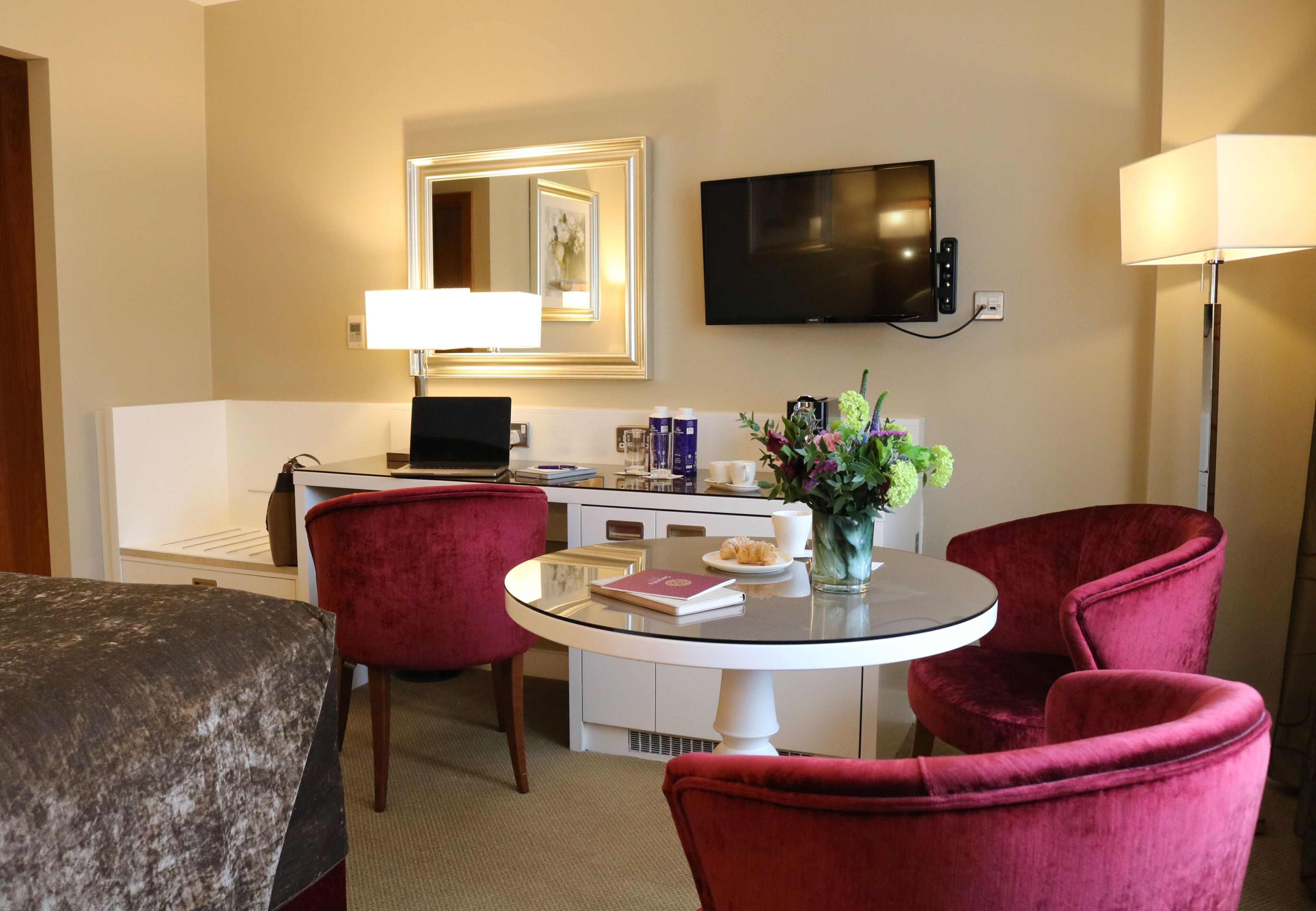 A hotel room with a bed, a desk with a computer, a TV, a lamp, a round table with chairs, and a vase with flowers.