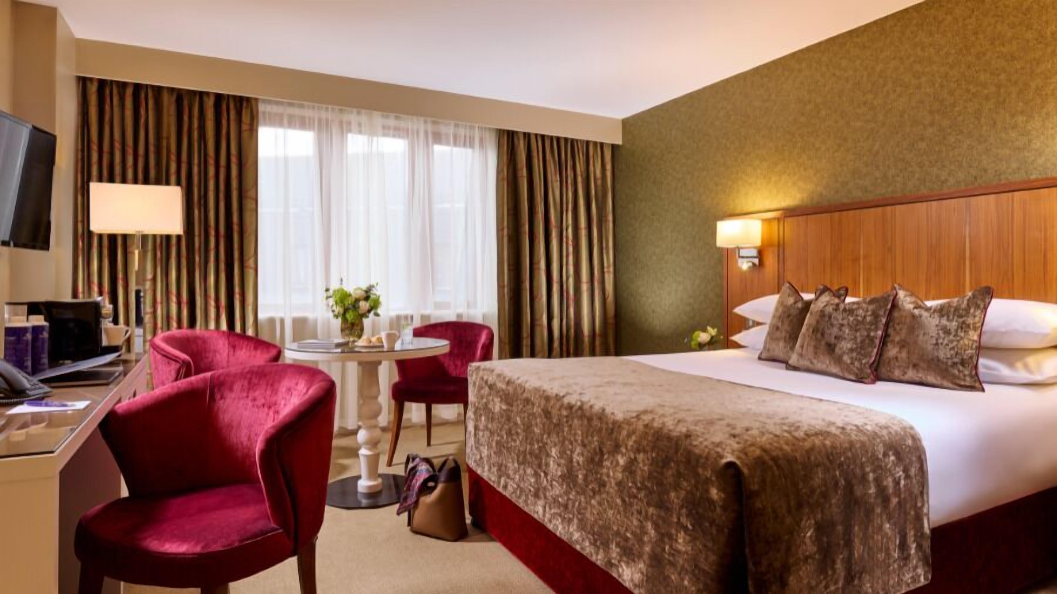 A hotel room with a bed, a desk with a computer, a chair, a small table with a vase, and curtains.