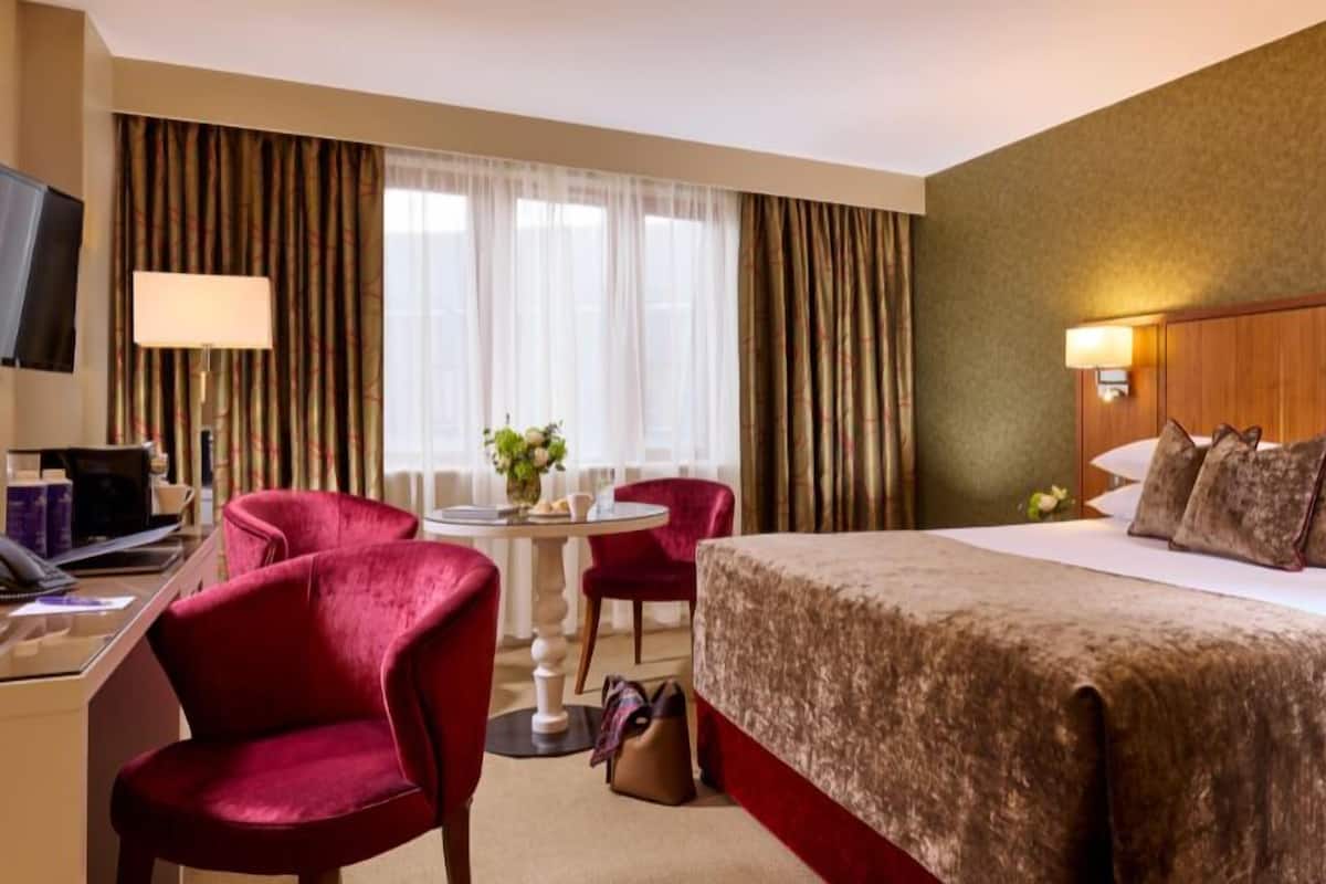 Habitación de hotel con una cama, un escritorio con computadora, una silla, una mesita con un florero y cortinas.