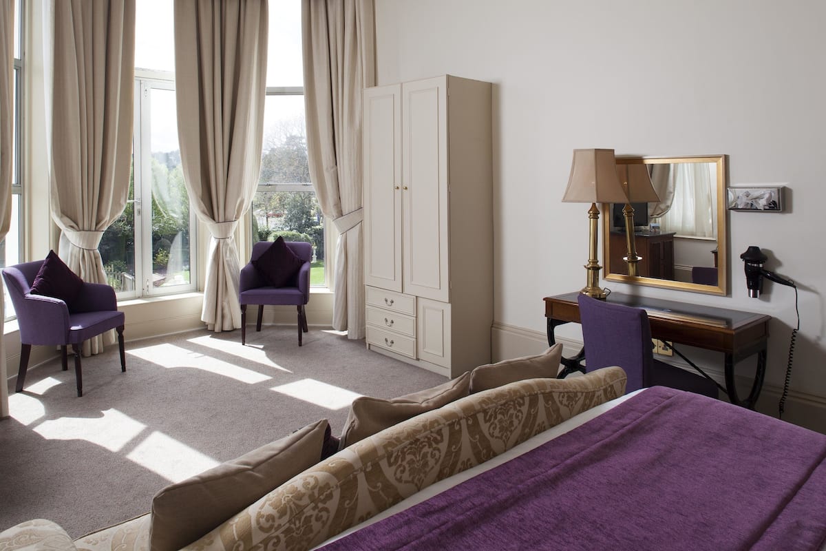 A bedroom with a bed, two chairs, a desk, a wardrobe, and a window with curtains.