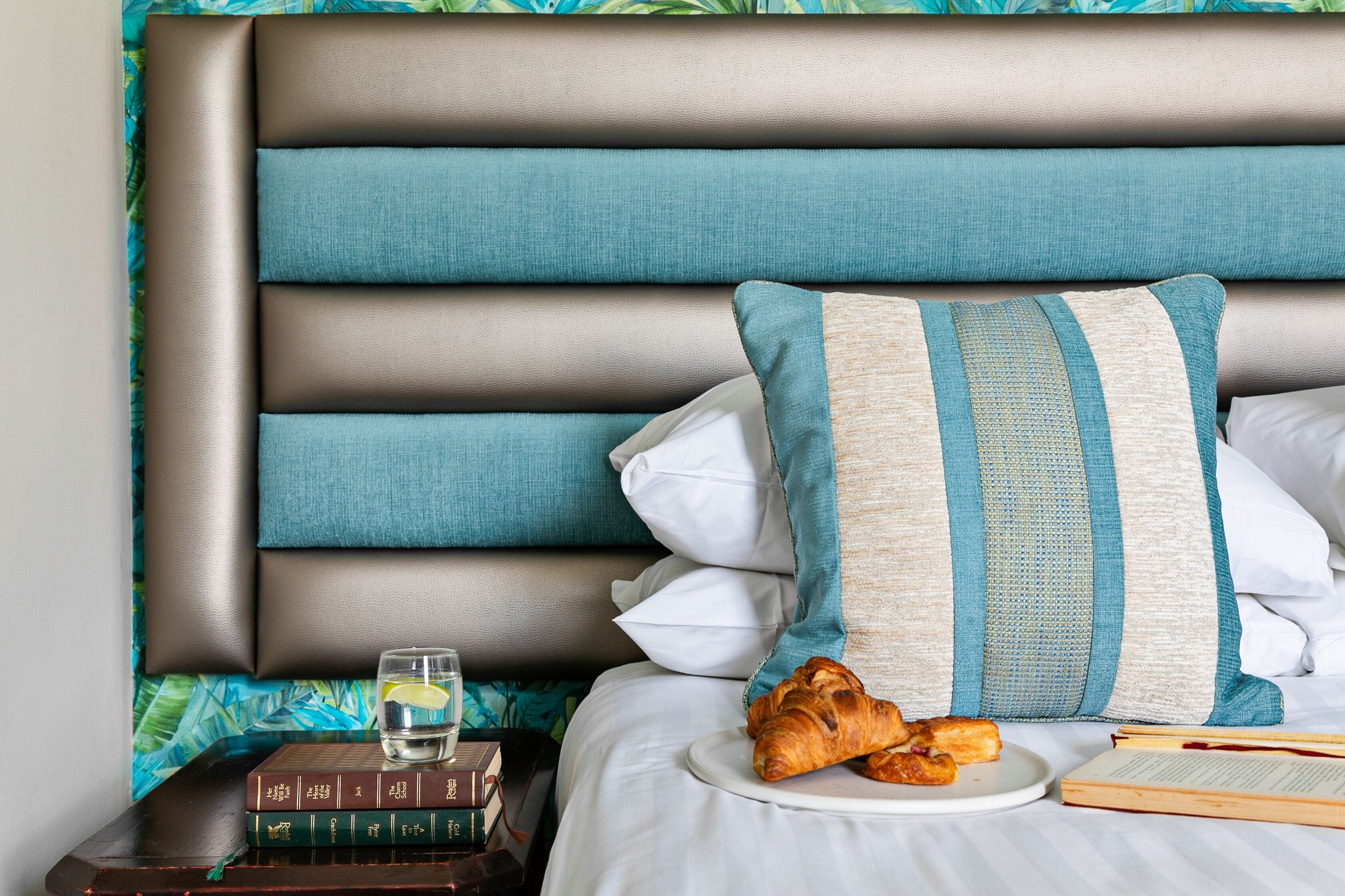 A neatly made bed with a striped pillow, a plate with croissants, a glass of water, and a book on a bedside table.