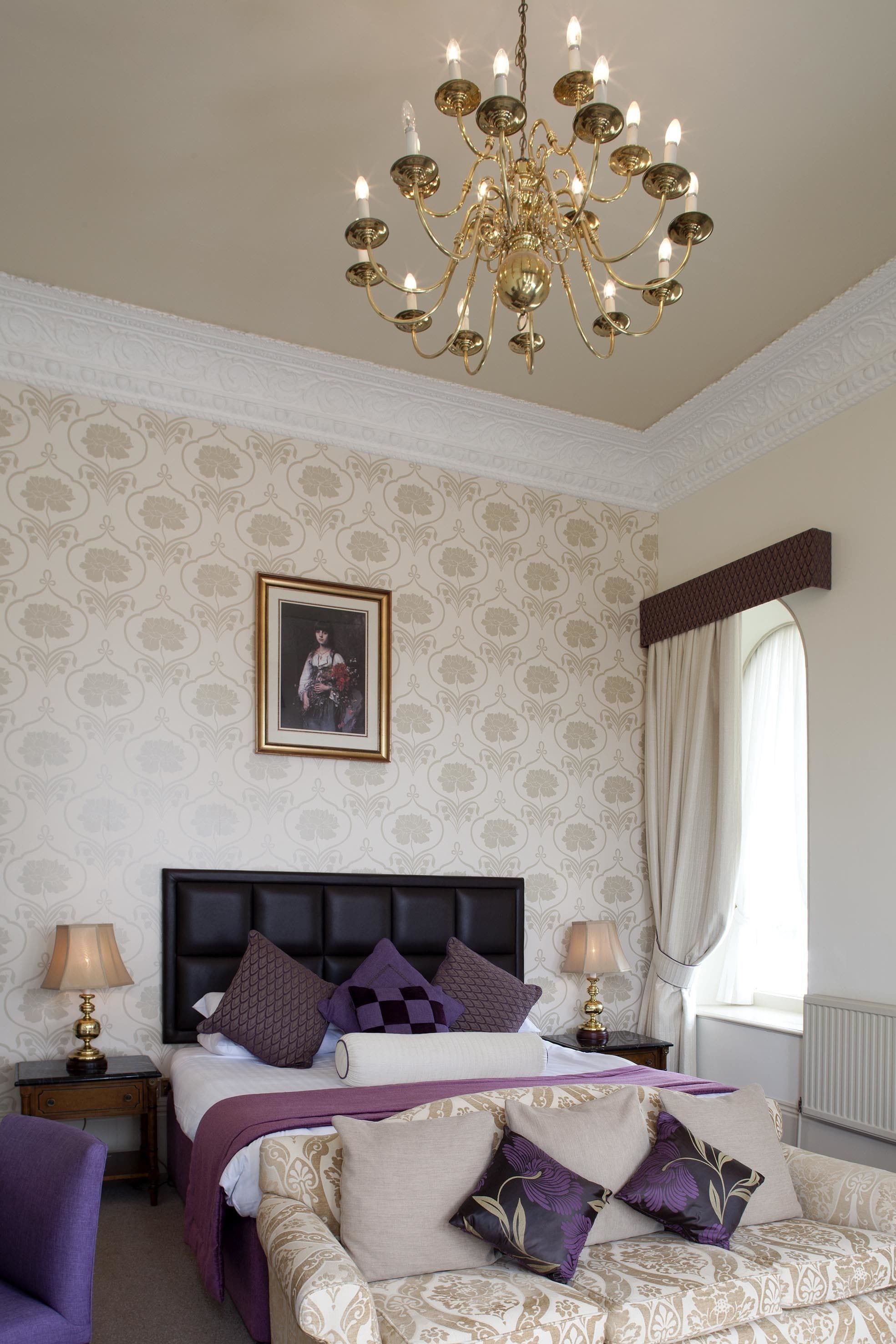 A bedroom with a large bed, a chandelier, a framed picture on the wall, and a window with curtains.