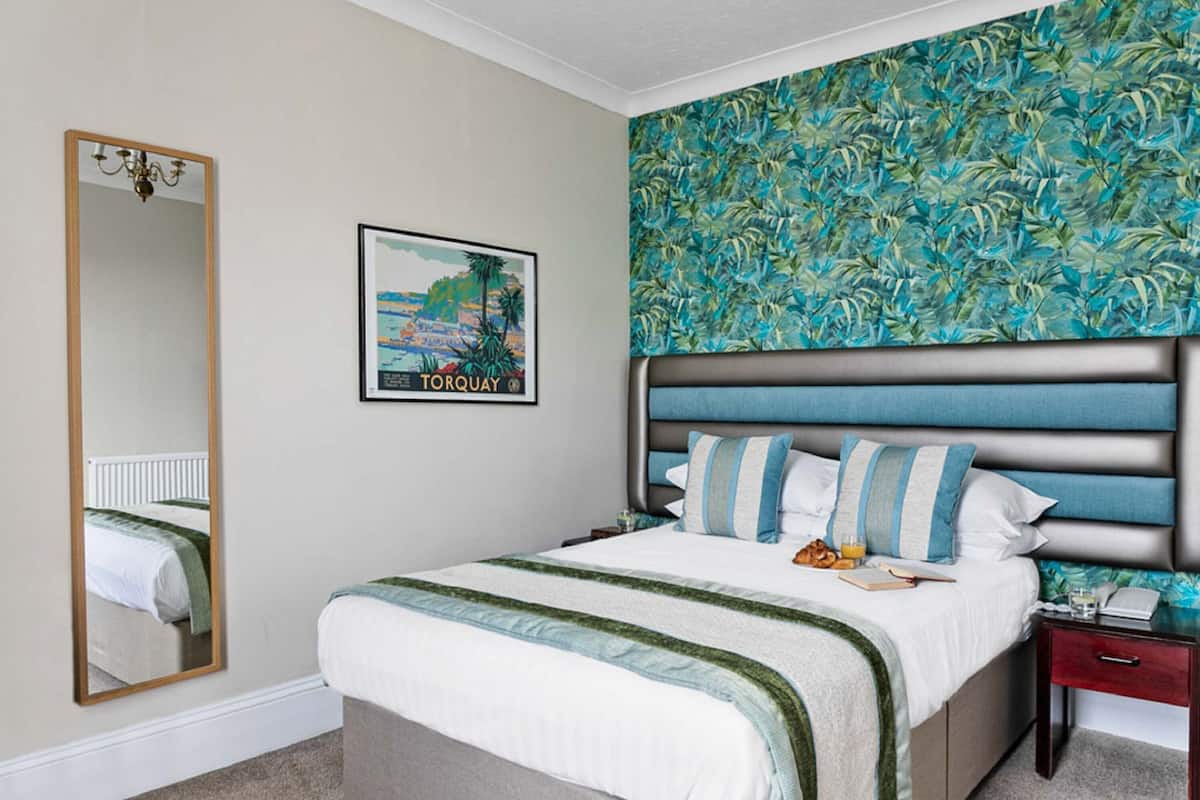 A bedroom with a bed, a nightstand, a mirror, and a wall with a tropical wallpaper.