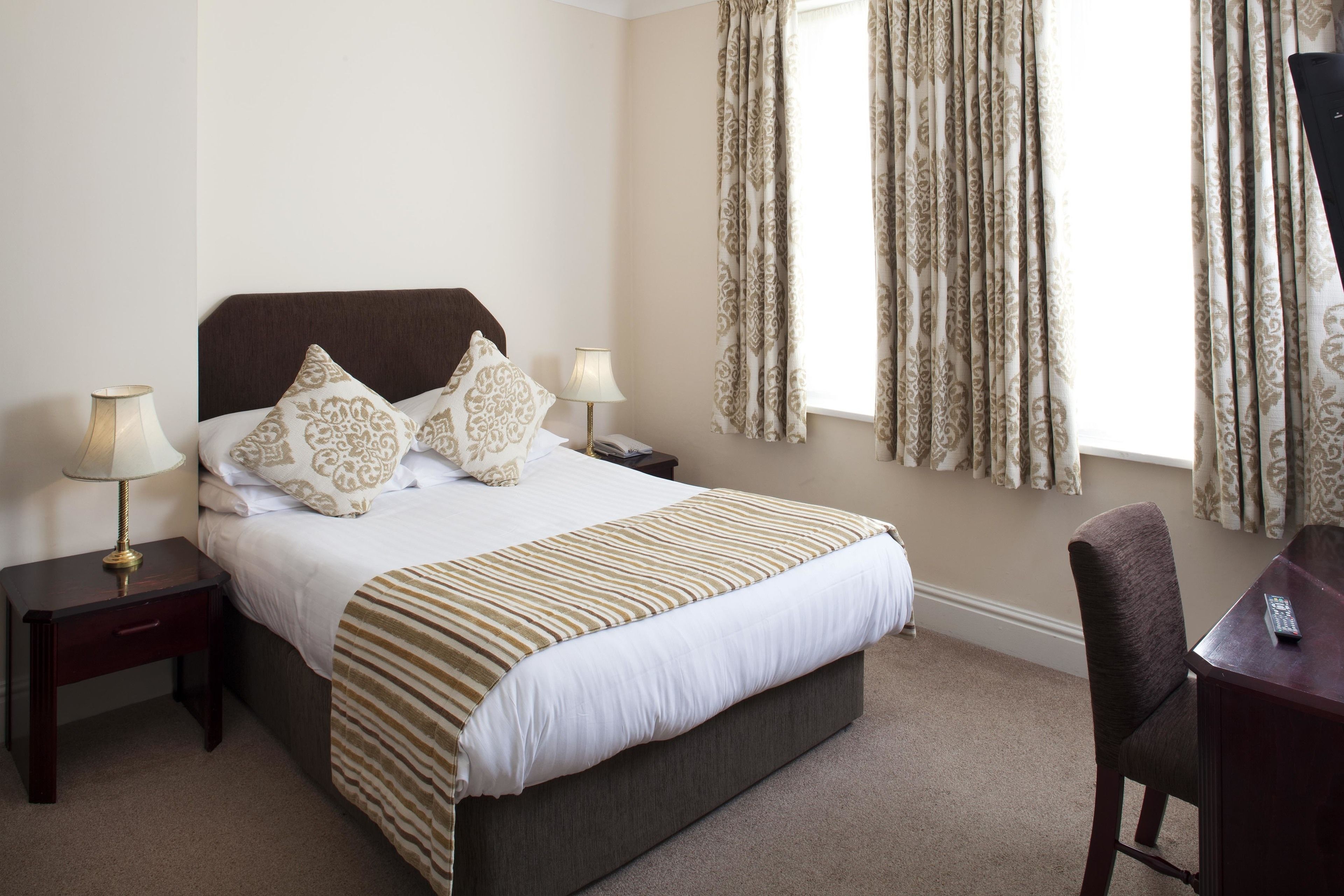 A neatly made bed with a striped bedspread, two bedside tables with lamps, a chair, and a window with curtains.