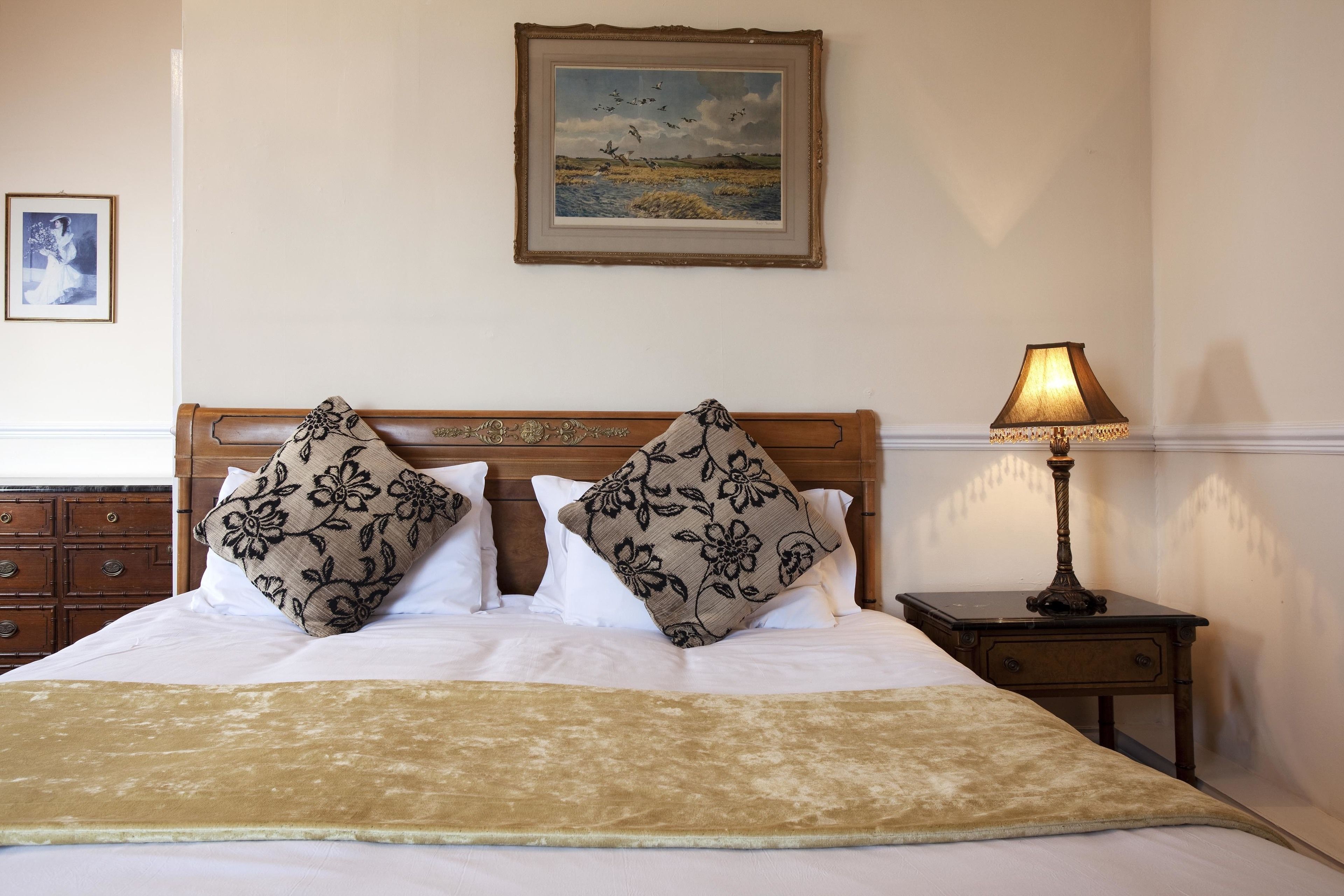 Una cama bien hecha con almohadas estampadas con motivos florales, una mesita de noche con una lámpara y un cuadro con un paisaje en la pared.