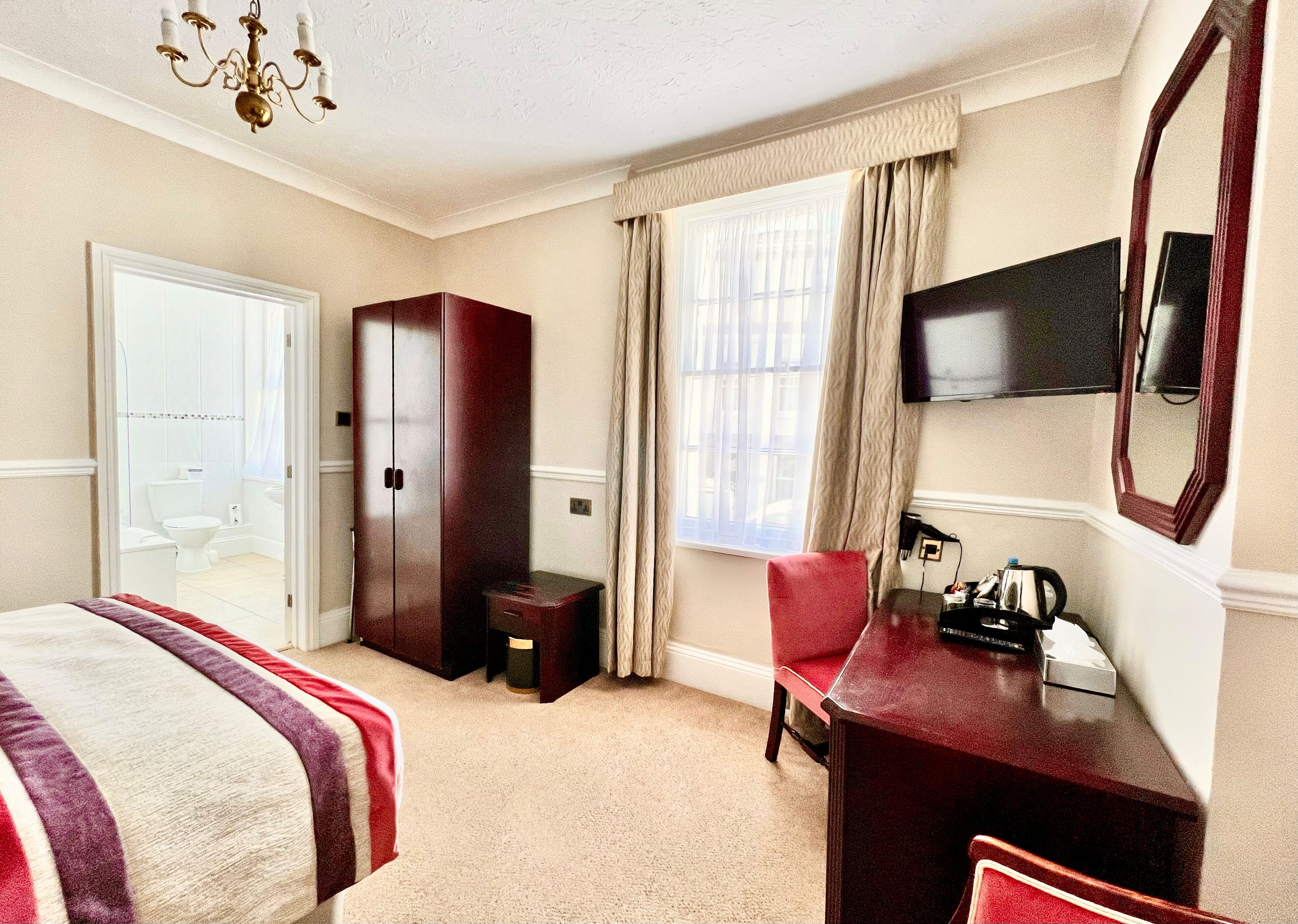 A hotel room with a bed, a desk, a chair, a wardrobe, a television, and a window with curtains.