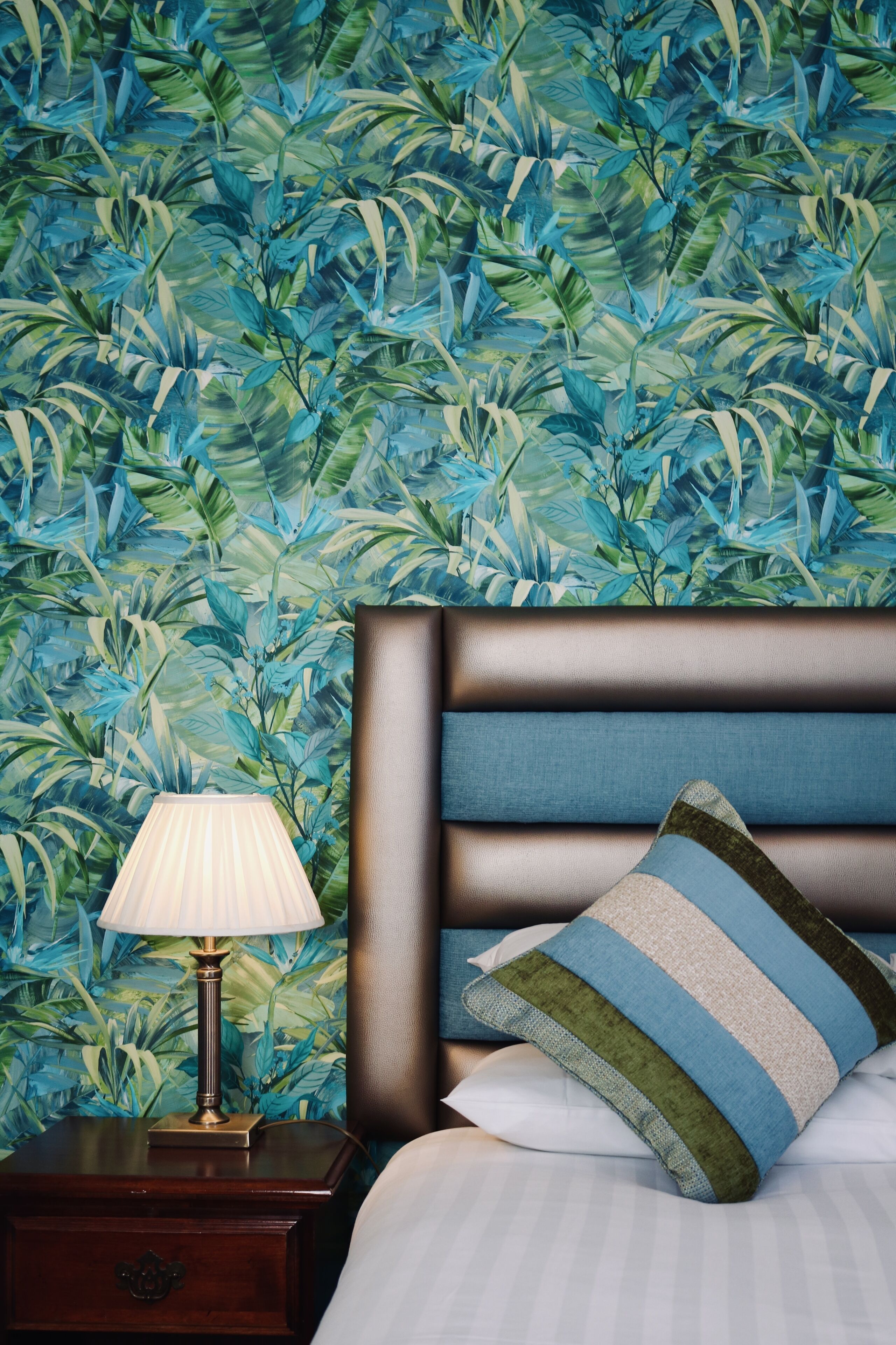 Une chambre à coucher comprenant un lit, une table de chevet, une lampe et un mur orné d’un motif de feuilles tropicales.