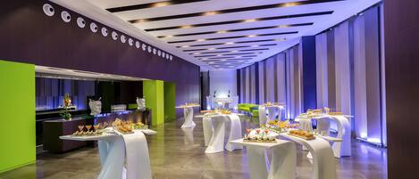 A modern hotel lobby with a long reception desk, multiple tables with white tablecloths, and a bright green accent wall.