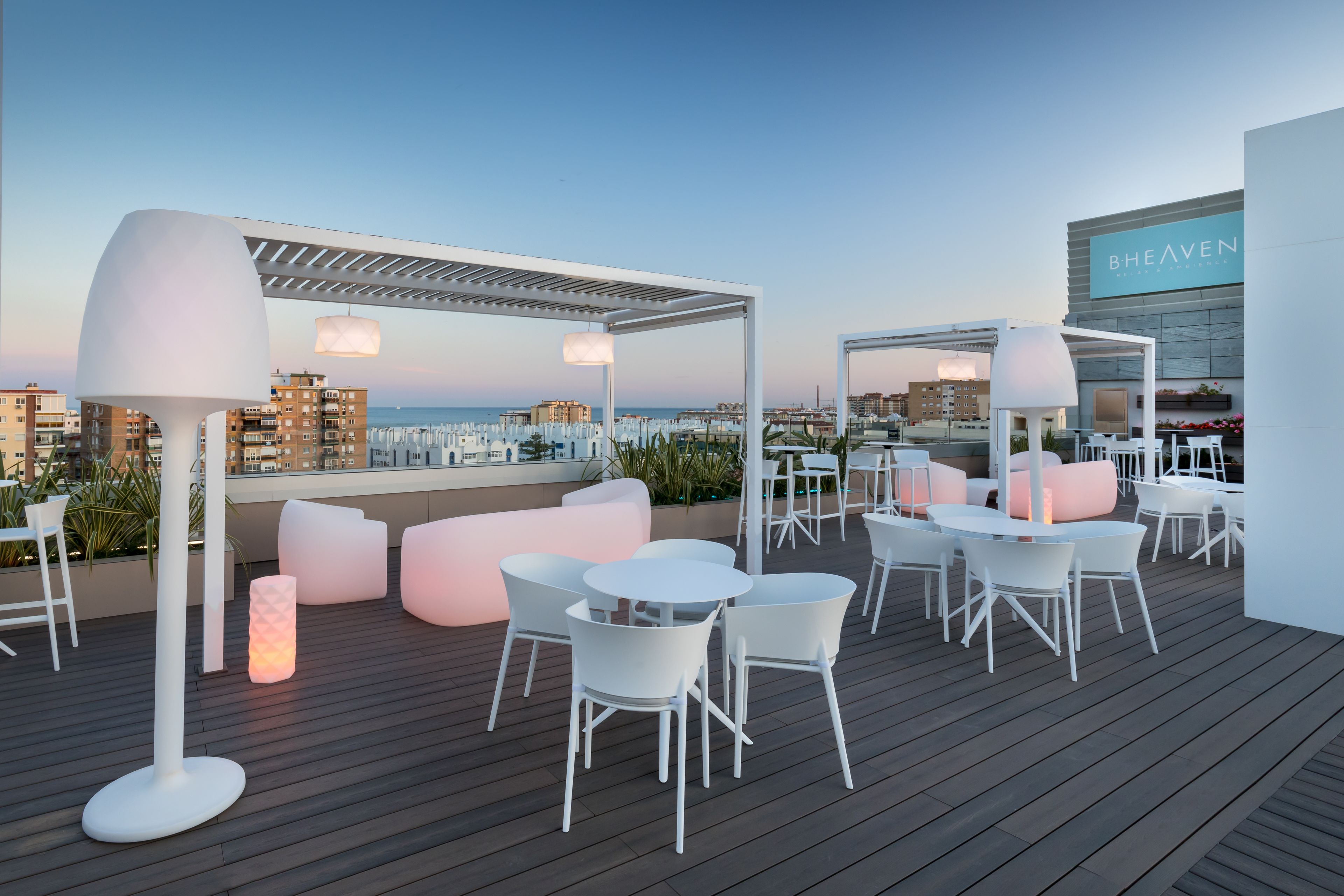 Una terraza en la azotea con mobiliario blanco, incluyendo mesas y sillas, bajo una pérgola blanca.