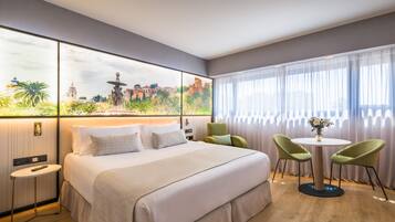 A hotel room with a bed, a small table, two chairs, a painting on the wall, and a window with curtains.