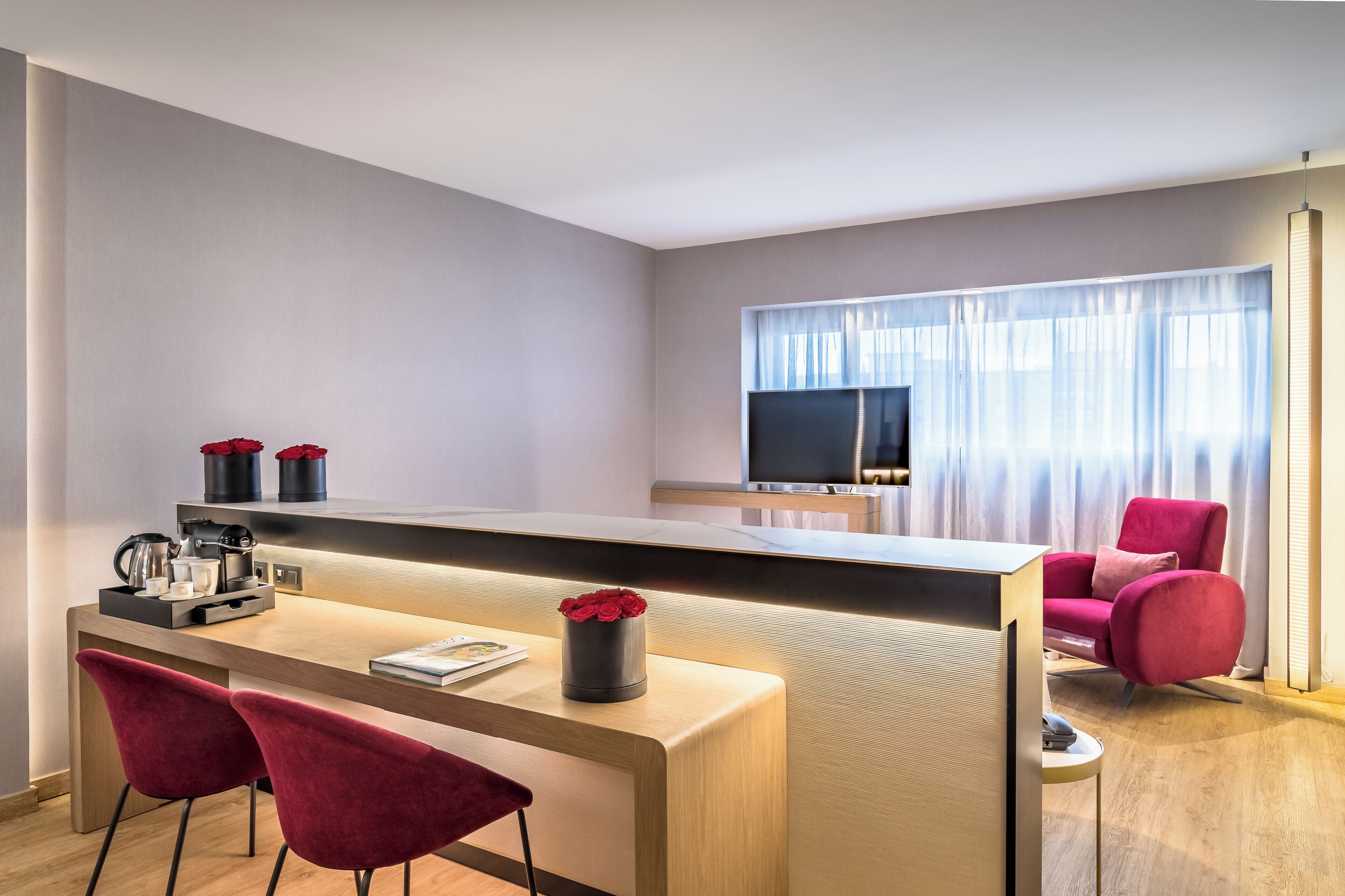 A modern hotel room with a wooden desk, two red chairs, a TV, and a large window with curtains.