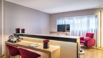 A modern hotel room with a wooden desk, two red chairs, a TV, and a large window with curtains.