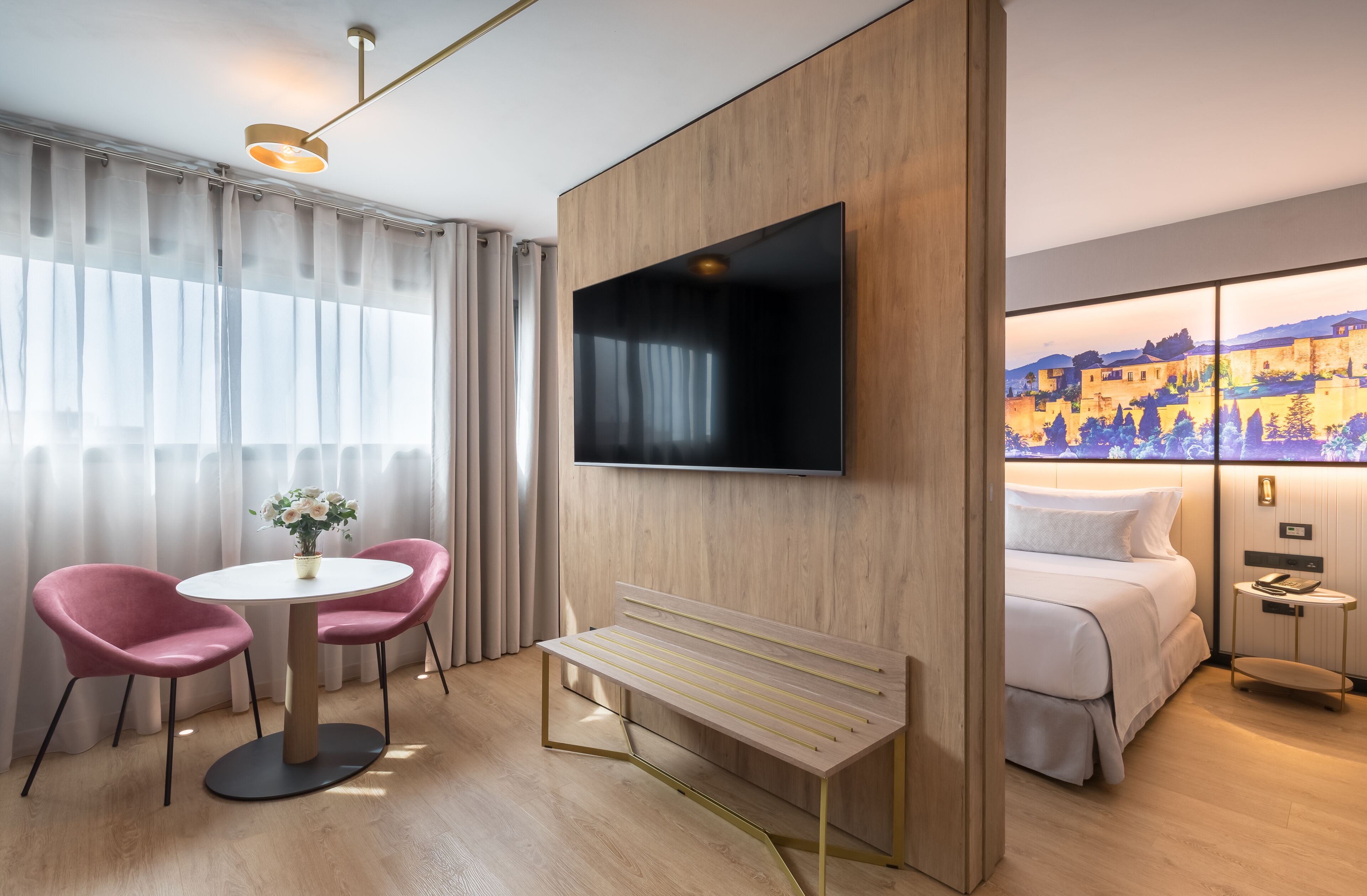 A hotel room with a bed, a TV mounted on the wall, a wooden bench, a round table with two chairs, and a view of a cityscape through the window.