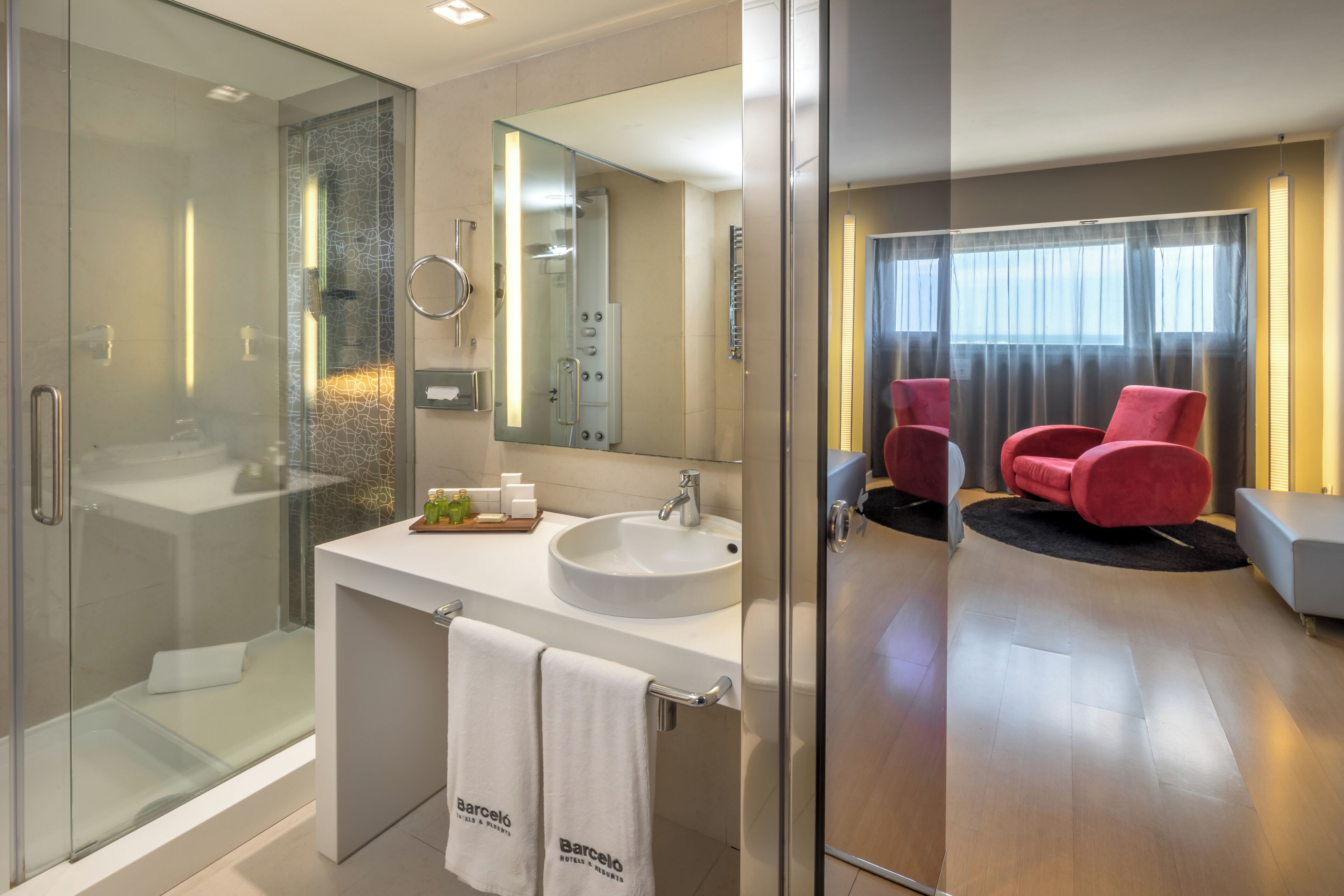 Un baño moderno con ducha de vidrio, un lavabo blanco y un espejo grande. En la zona de estar contigua hay dos sillas rojas.