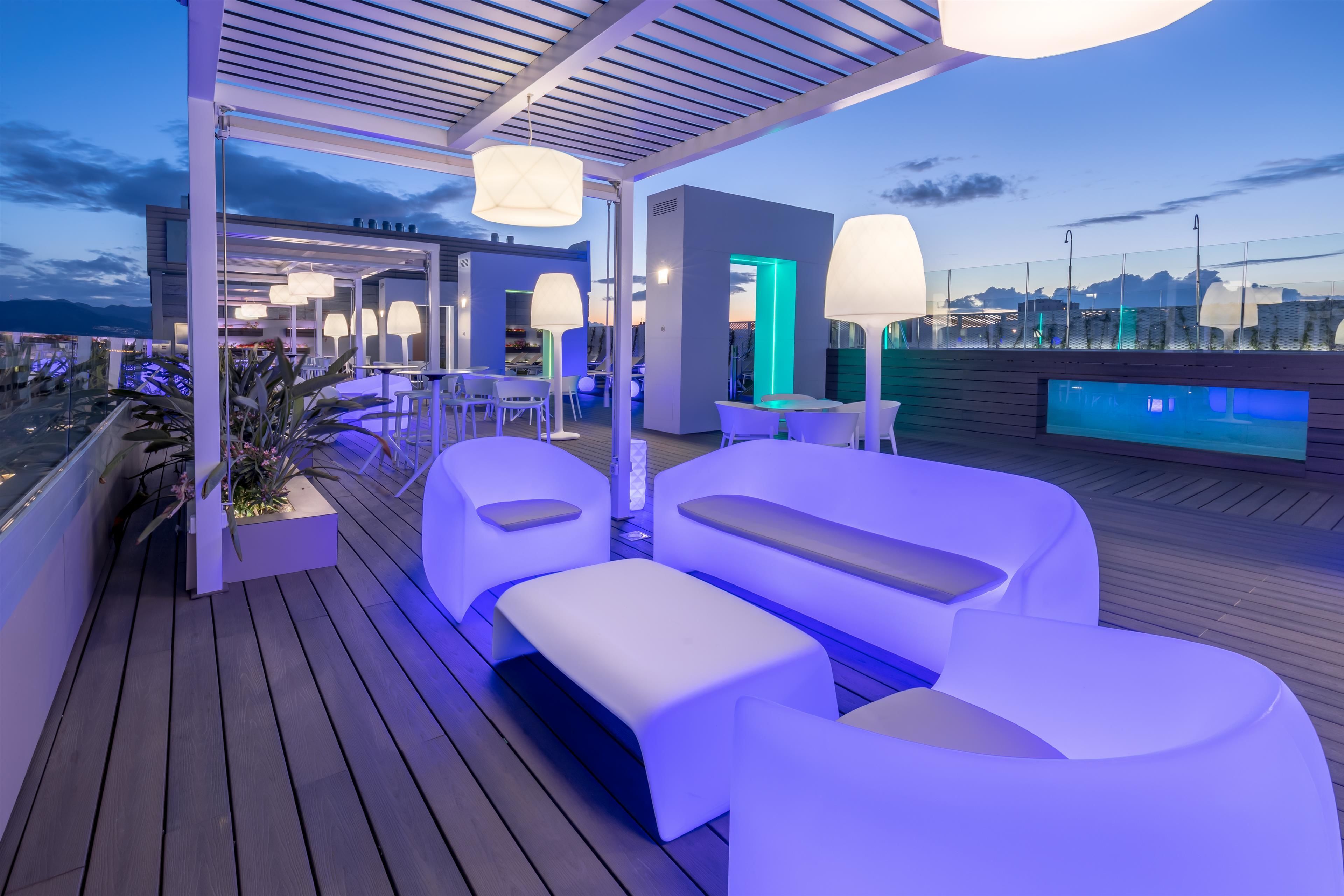 A modern outdoor lounge area with white furniture and ambient lighting, overlooking a cityscape.