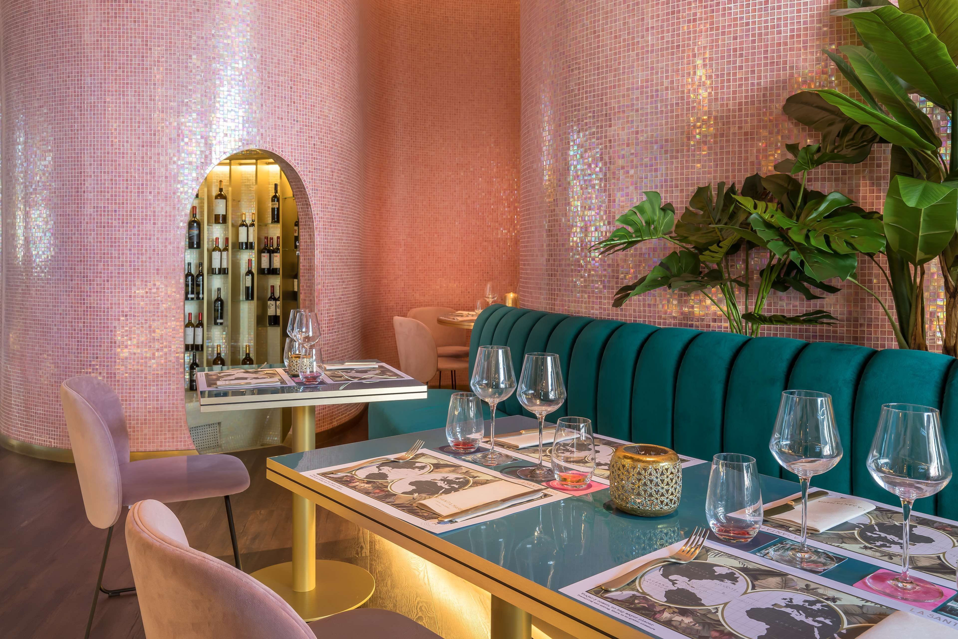 En restaurant med lyserøde mosaikvægge, lyseblå polstrede siddepladser og et vinskab, der ses igennem en bue.