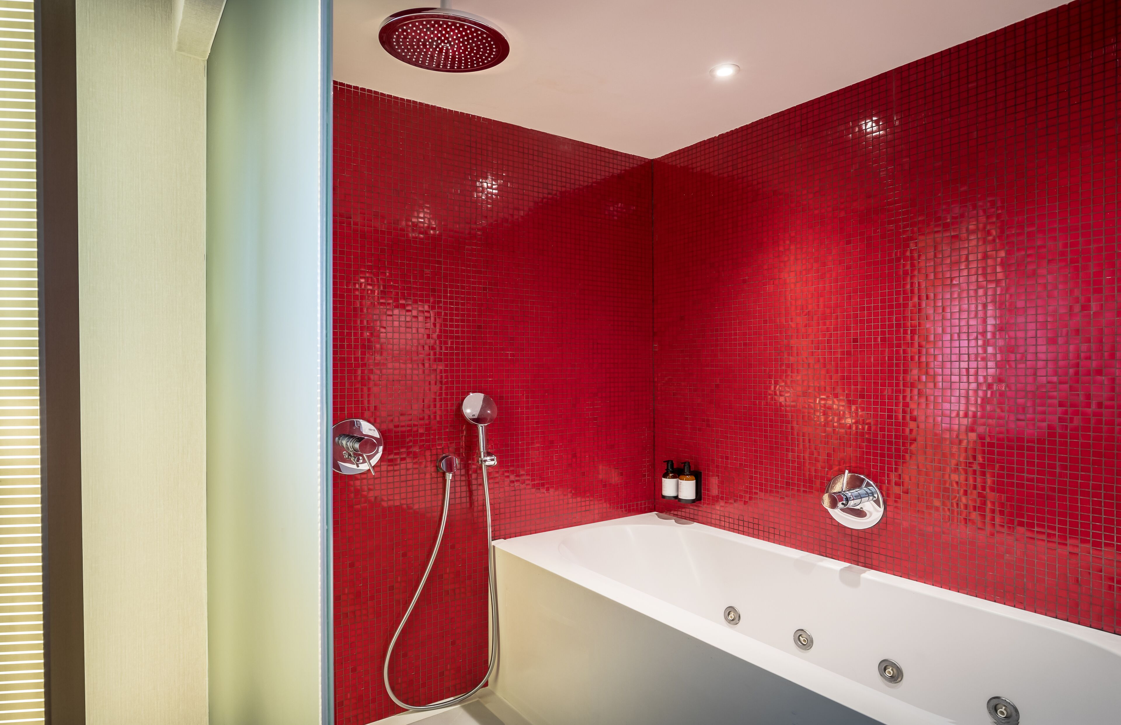 A modern bathroom with a red tiled wall, a white bathtub, and a shower with a handheld showerhead.