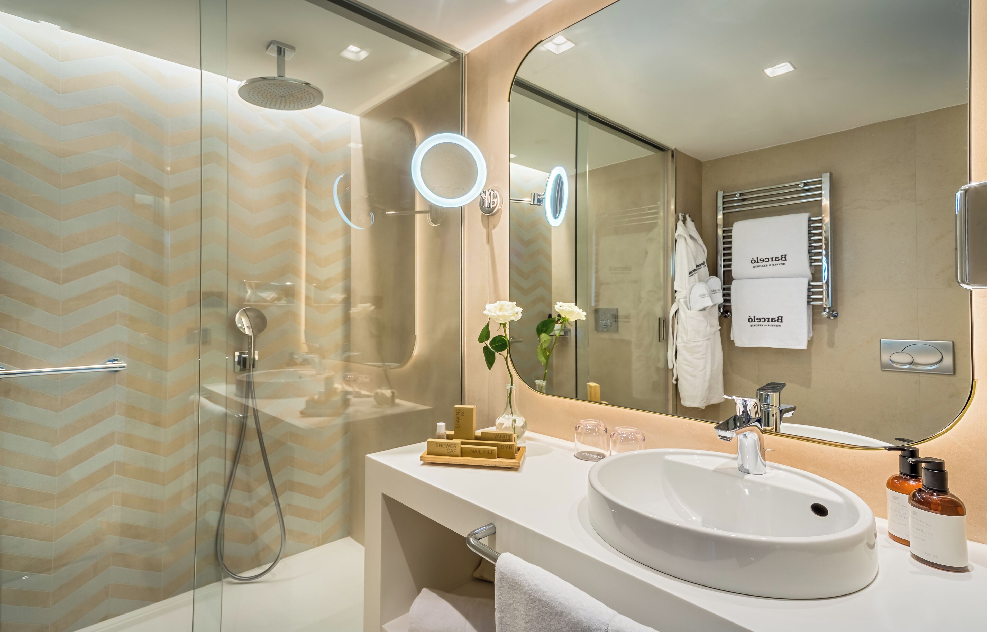 A modern bathroom with a glass shower enclosure, a large mirror with a ring light, a white sink, and a towel rack.