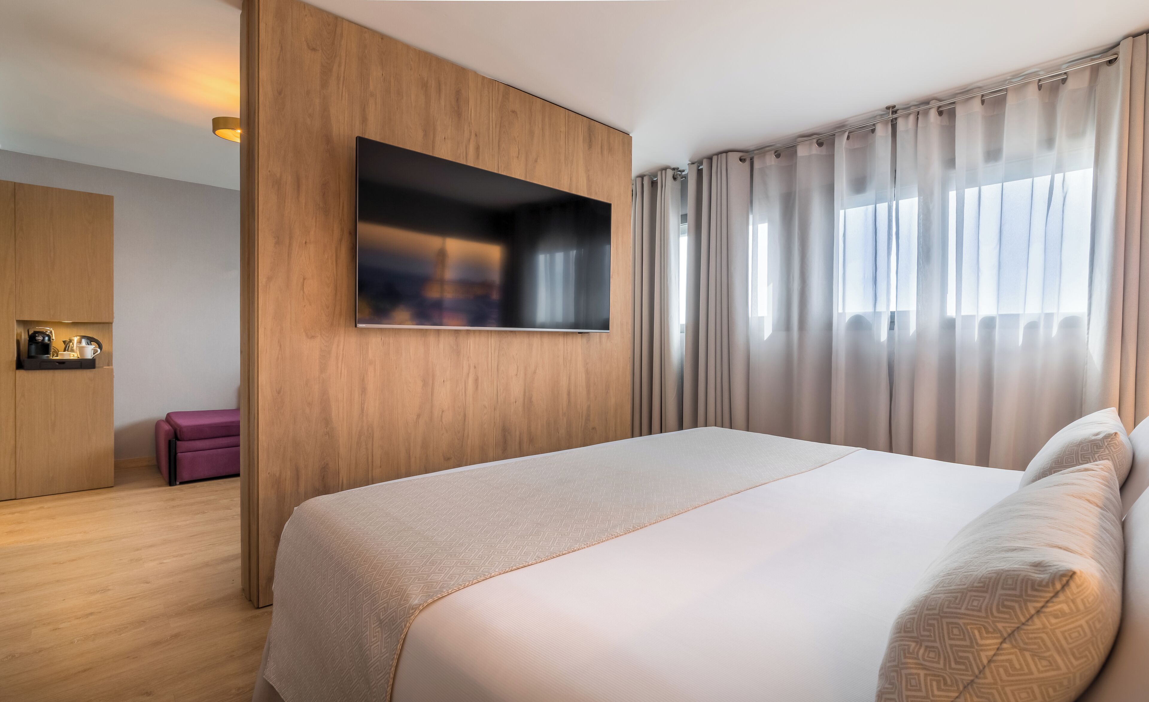 A modern hotel room with a large bed, a flat-screen TV mounted on a wooden panel, a small seating area, and a window with curtains.
