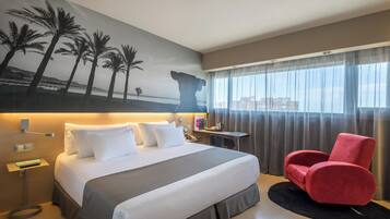Habitación de hotel con una cama grande, una silla roja, una mesita de noche y un cuadro de palmeras.