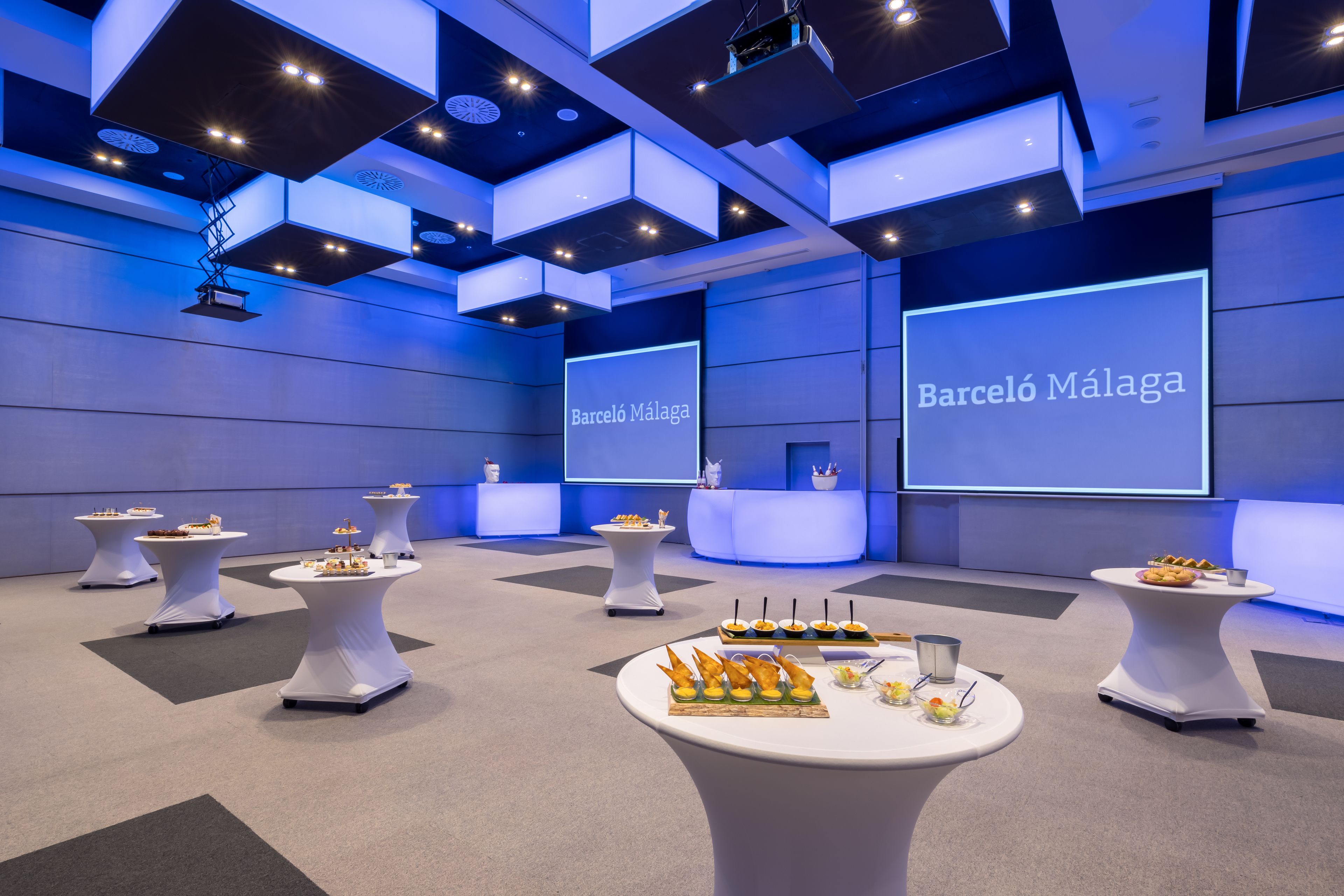A modern conference room with multiple tables displaying food and beverages, a large screen with the text "Barceló Málaga", and a stage area.