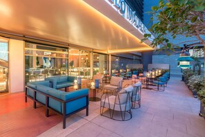 Un área de estar al aire libre con sofás azules y mesas, rodeada de paredes de vidrio y una fachada de edificio moderna.