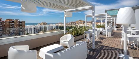 A rooftop terrace with white furniture, a pergola, and a view of the city and sea.