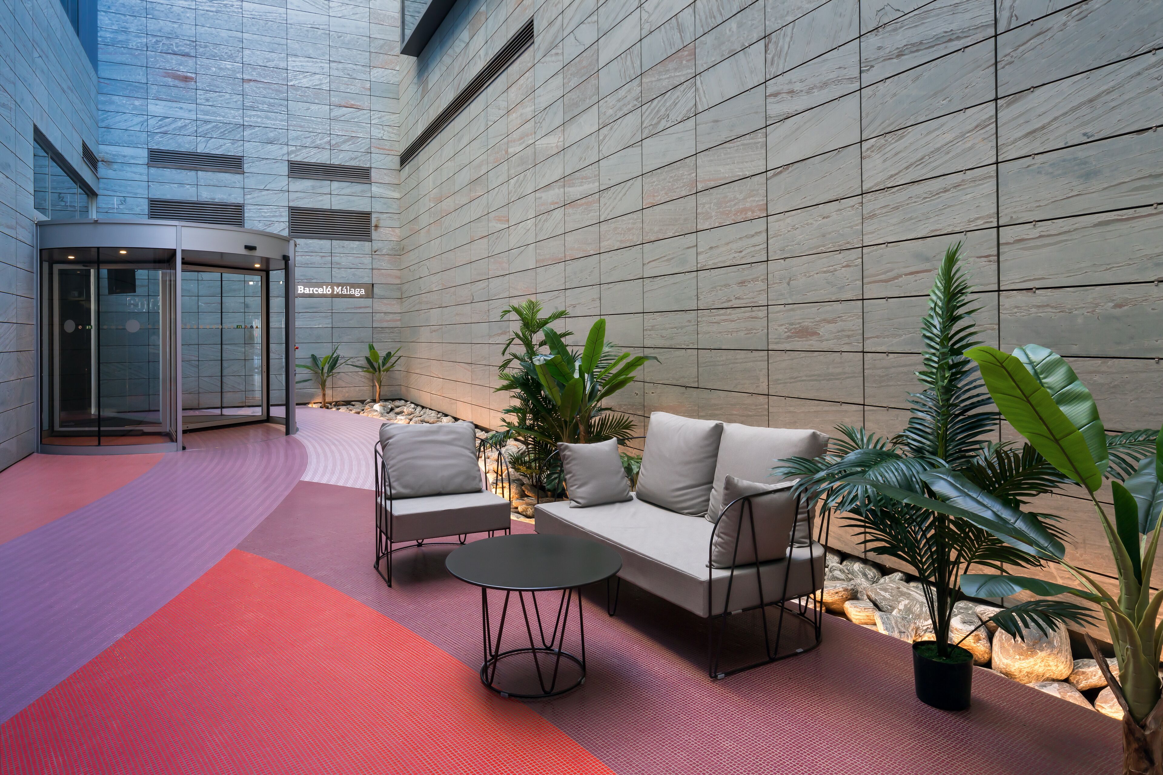 Um lobby moderno com entrada em vidro curvo, área de estar com sofá e poltronas, e plantas em vasos.
