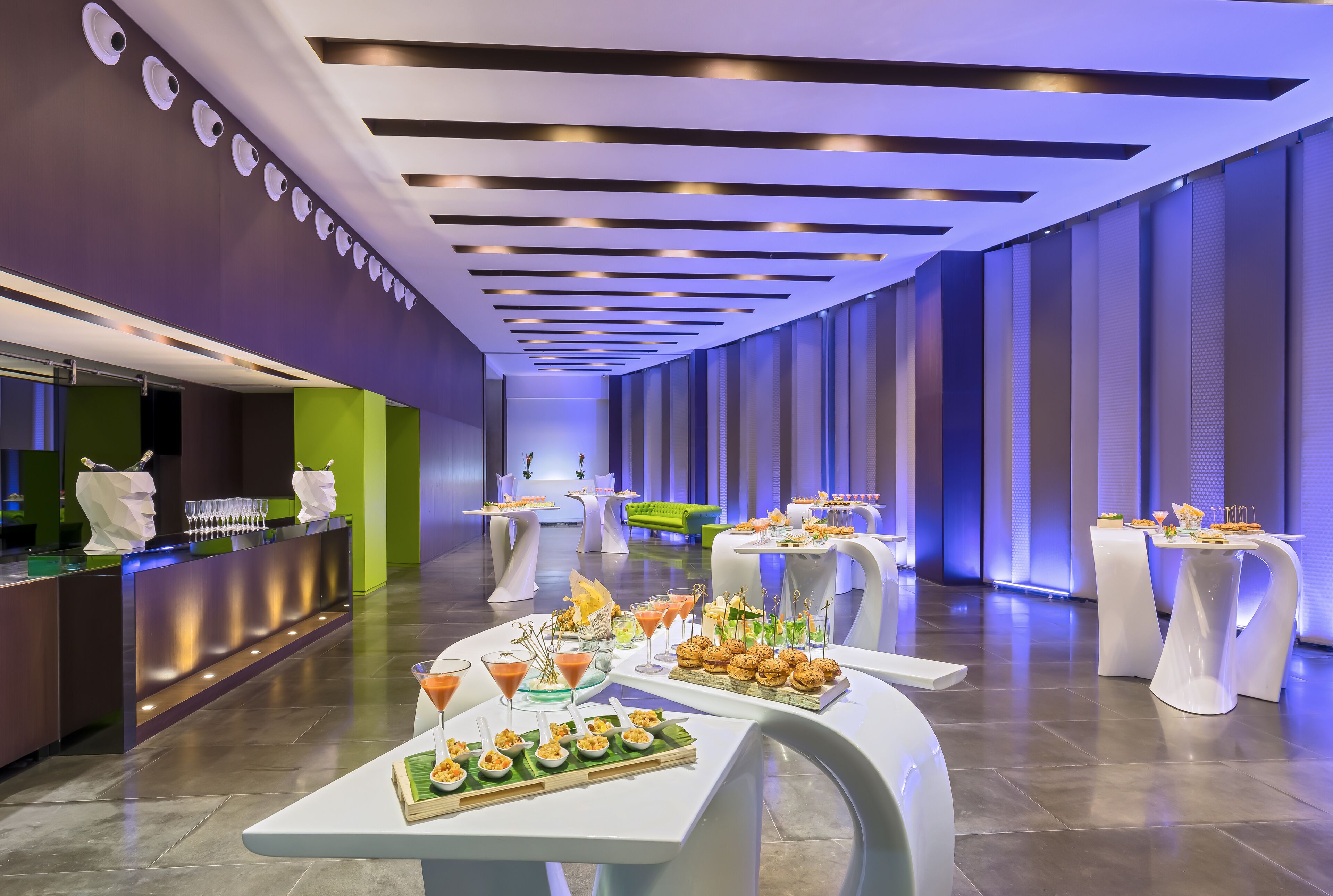 Un hall d’hôtel moderne avec un comptoir d’accueil, plusieurs tables où l’on peut prendre un repas ou boire un verre, et un canapé vert.