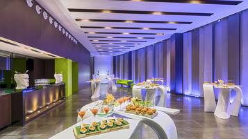 Un hall d’hôtel moderne avec un comptoir d’accueil, plusieurs tables où l’on peut prendre un repas ou boire un verre, et un canapé vert.