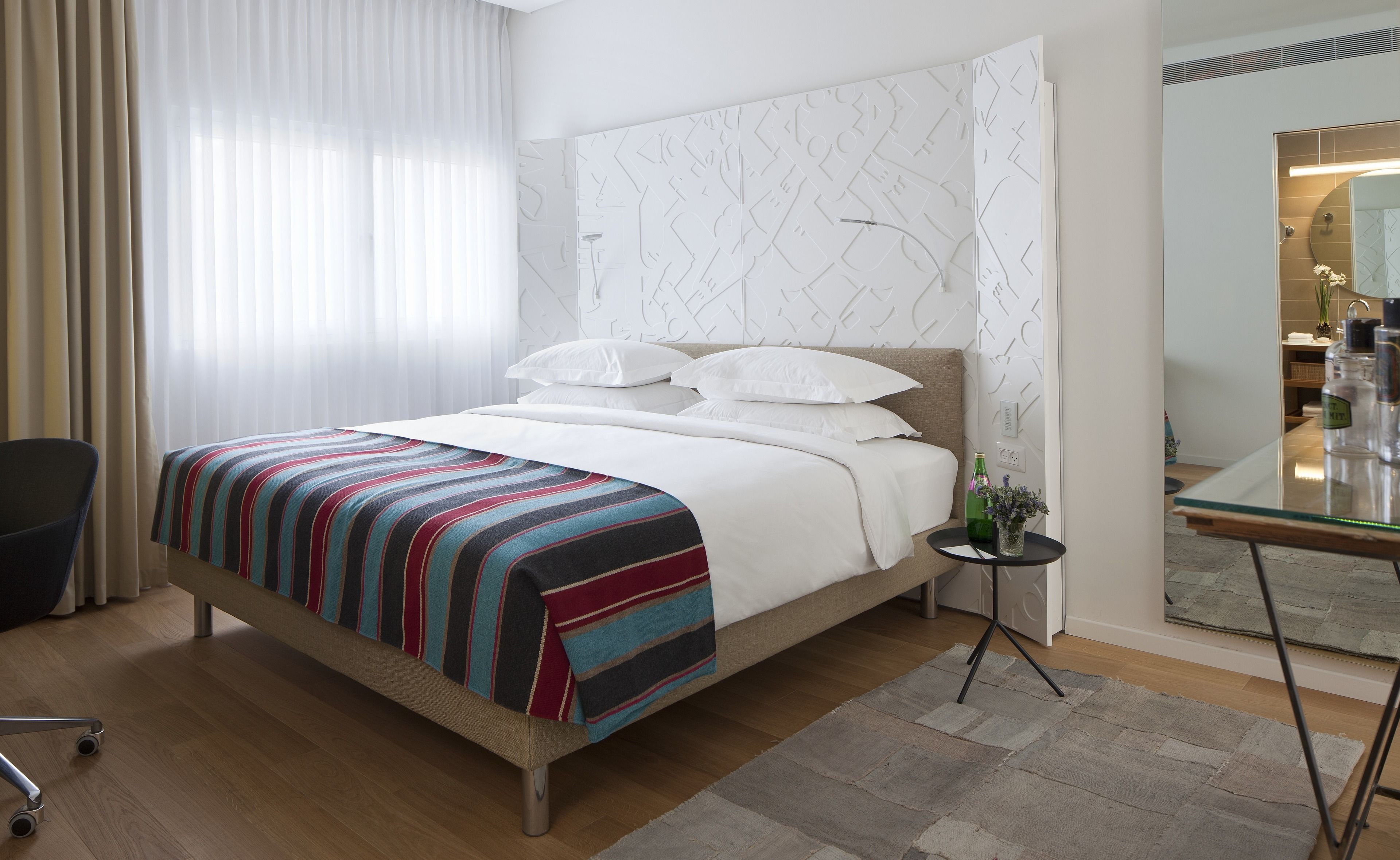 A modern hotel room with a bed, a small table with a vase, and a mirror.