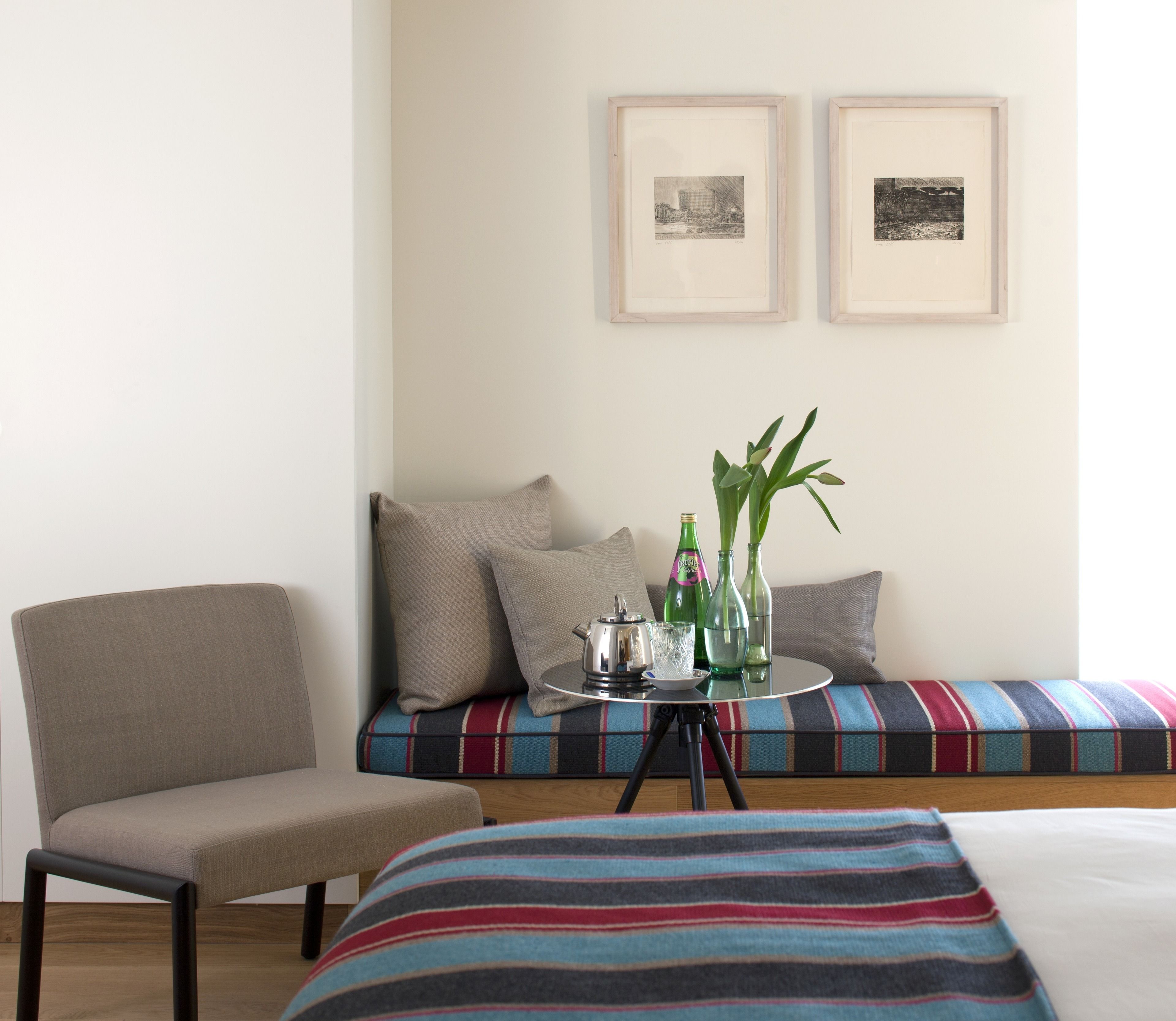 A room with a bed, a chair, a sofa with cushions, a small table with a teapot, glasses, and bottles, and two framed pictures on the wall.