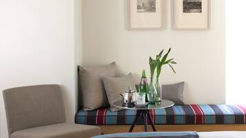A room with a bed, a chair, a sofa with cushions, a small table with a teapot, glasses, and bottles, and two framed pictures on the wall.