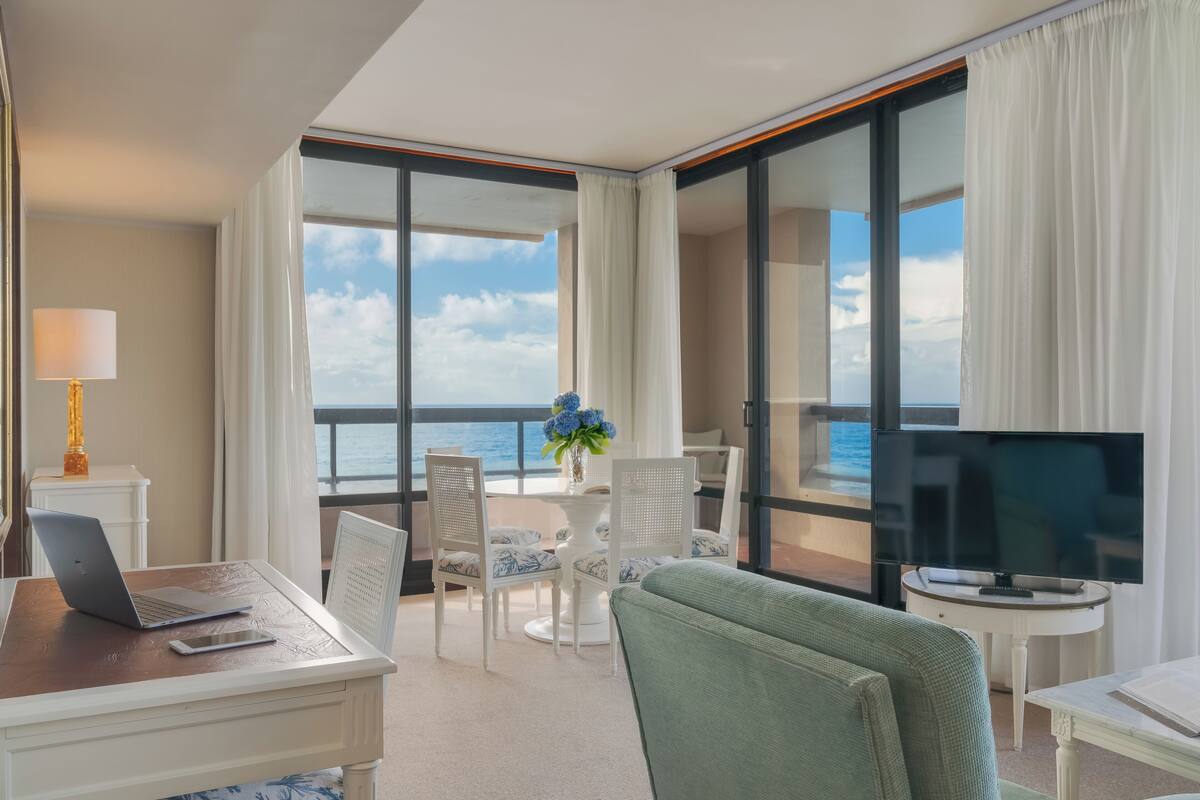 Ett hotellrum med ett skrivbord, en bärbar dator och en tv, stora fönster med utsikt över havet och en balkong med ett bord och stolar.